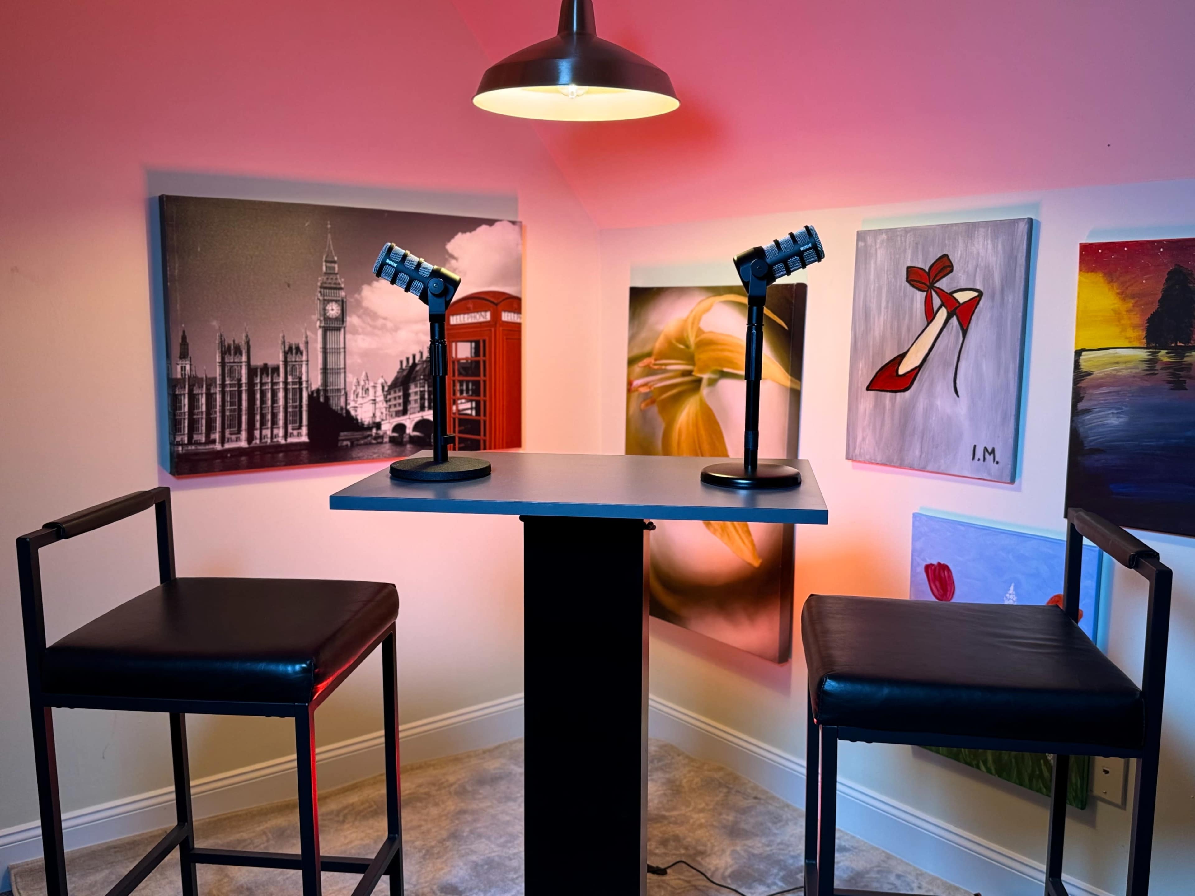 The image shows a small interview or podcast setup featuring two tall stools and a central table, illuminated by a pendant lamp, with various artworks displayed on the walls around it.