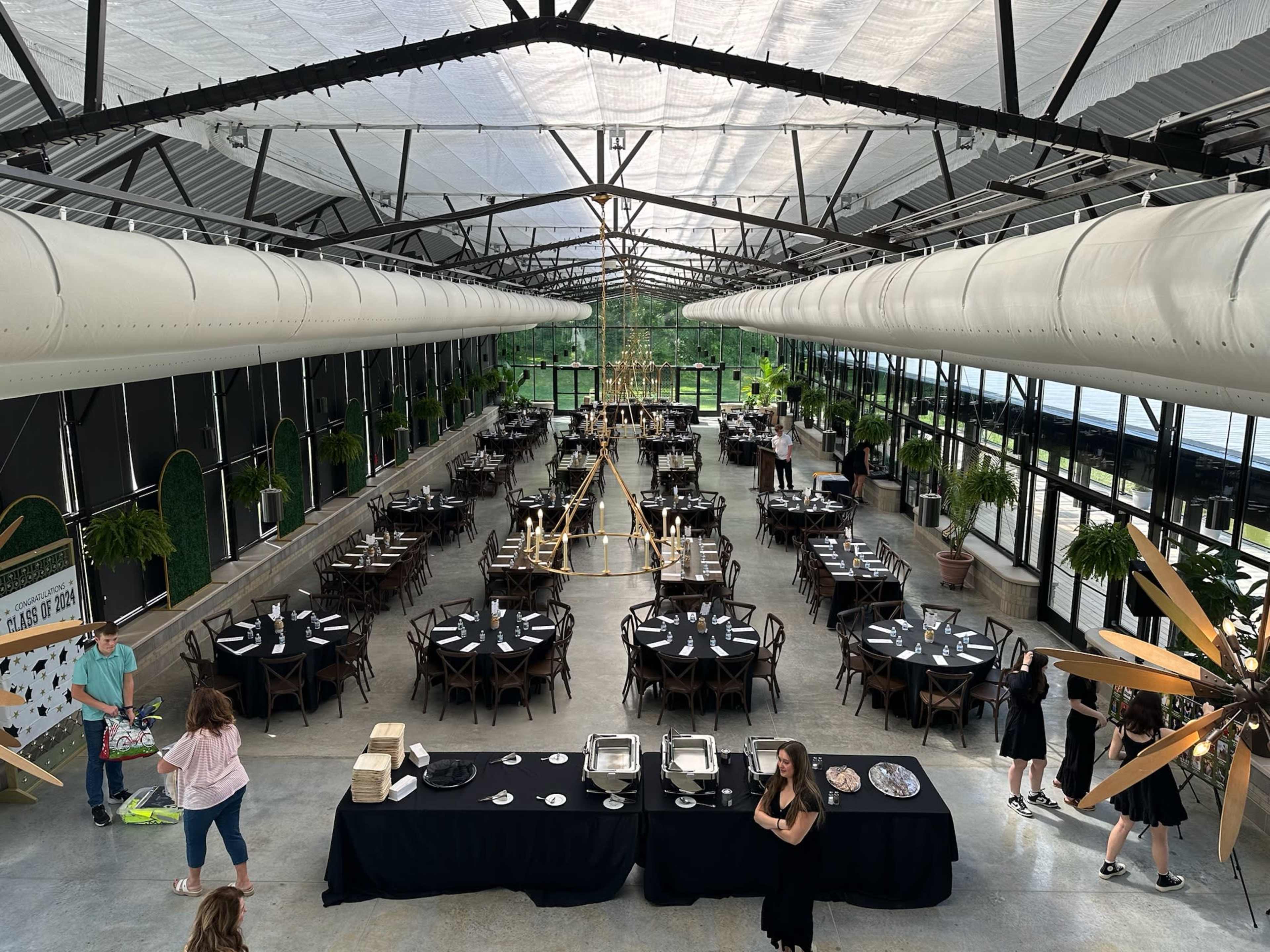 The image shows a large dining hall with multiple round tables set for a formal event, featuring a modern design and greenery along the windows.