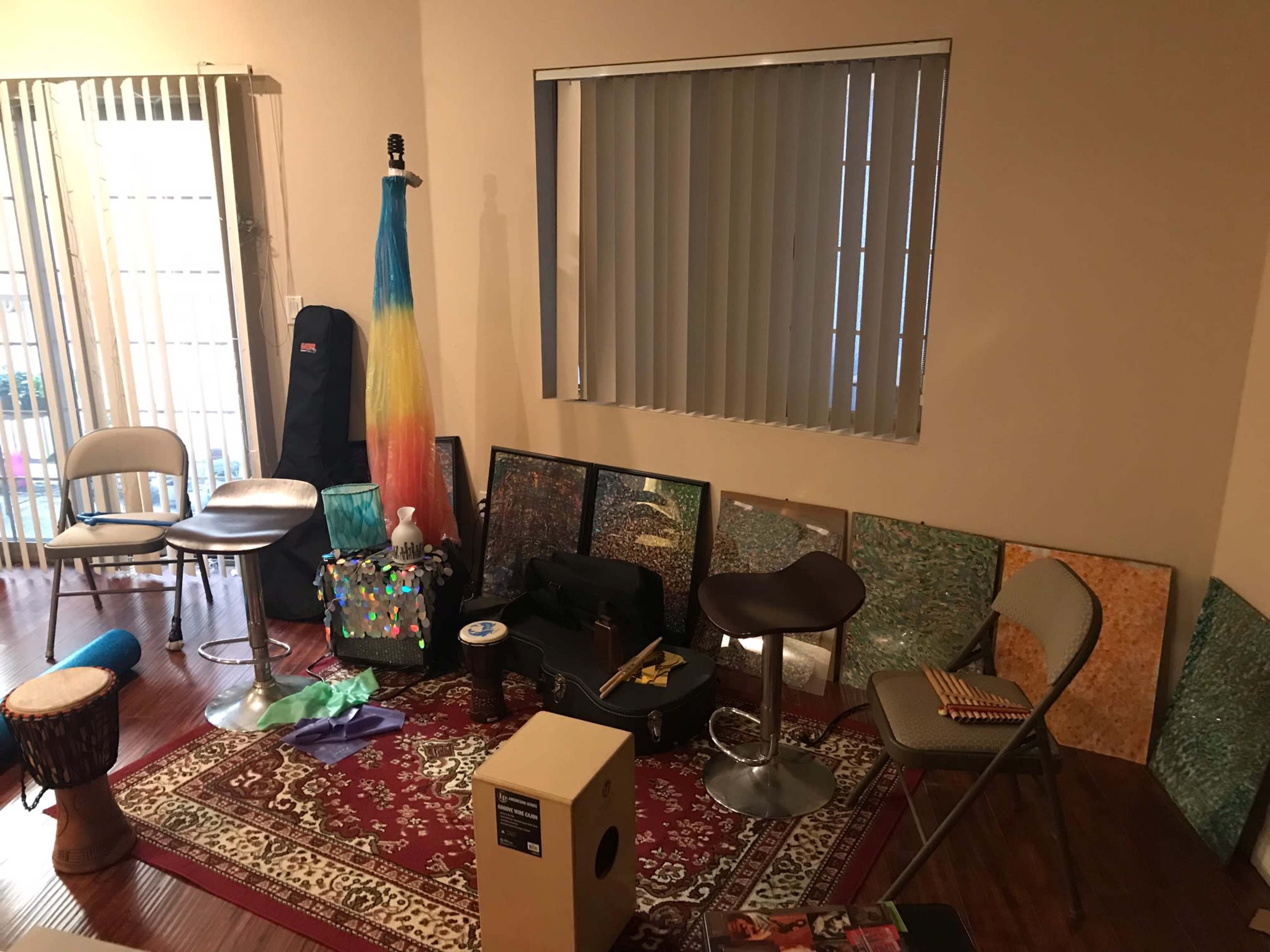 An assortment of musical instruments and artwork is arranged in a room with wooden flooring and large windows covered by vertical blinds.