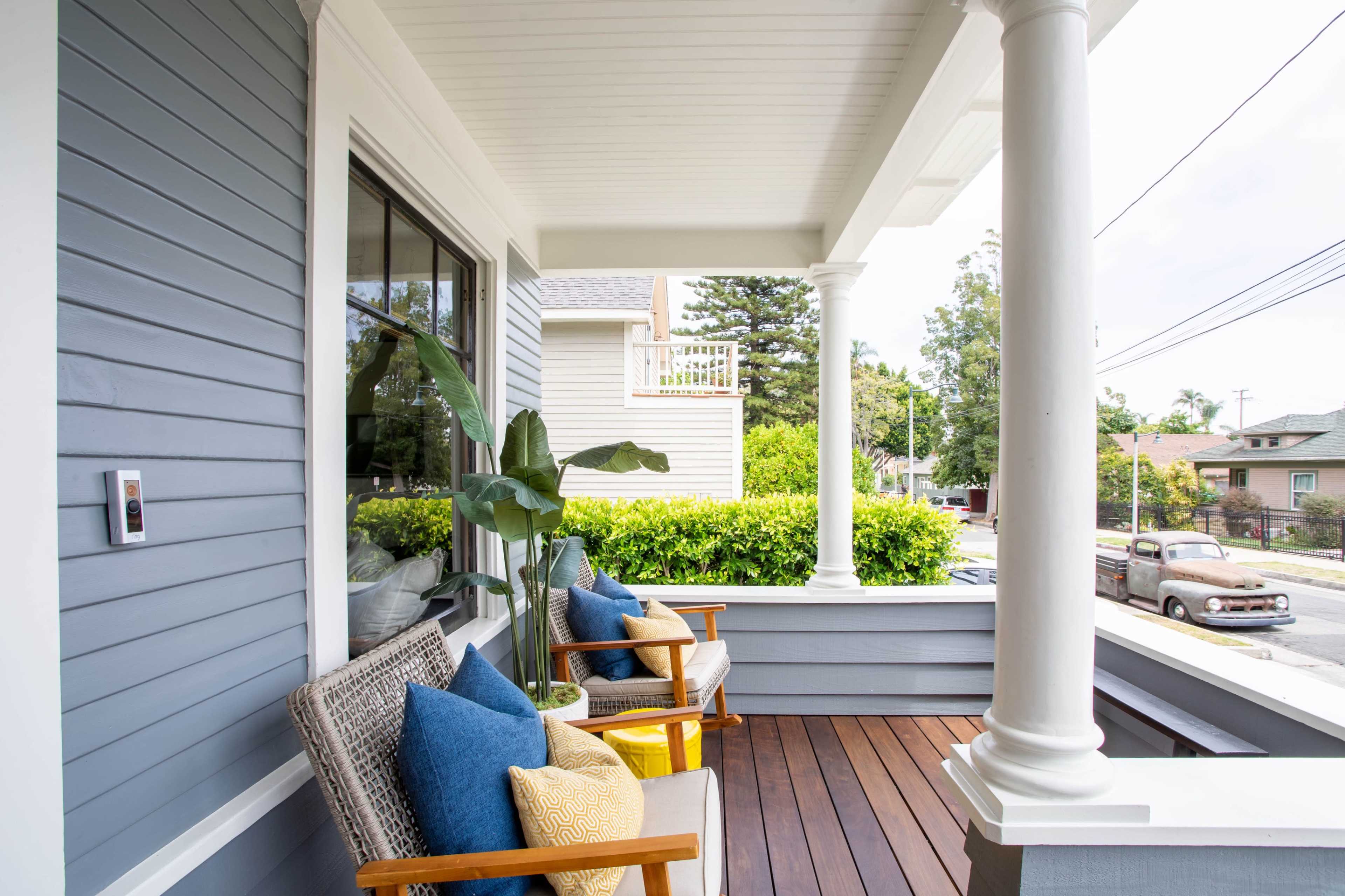 A porch with two wooden chairs adorned with colorful pillows, facing a lush green hedge and a vintage car parked nearby.
