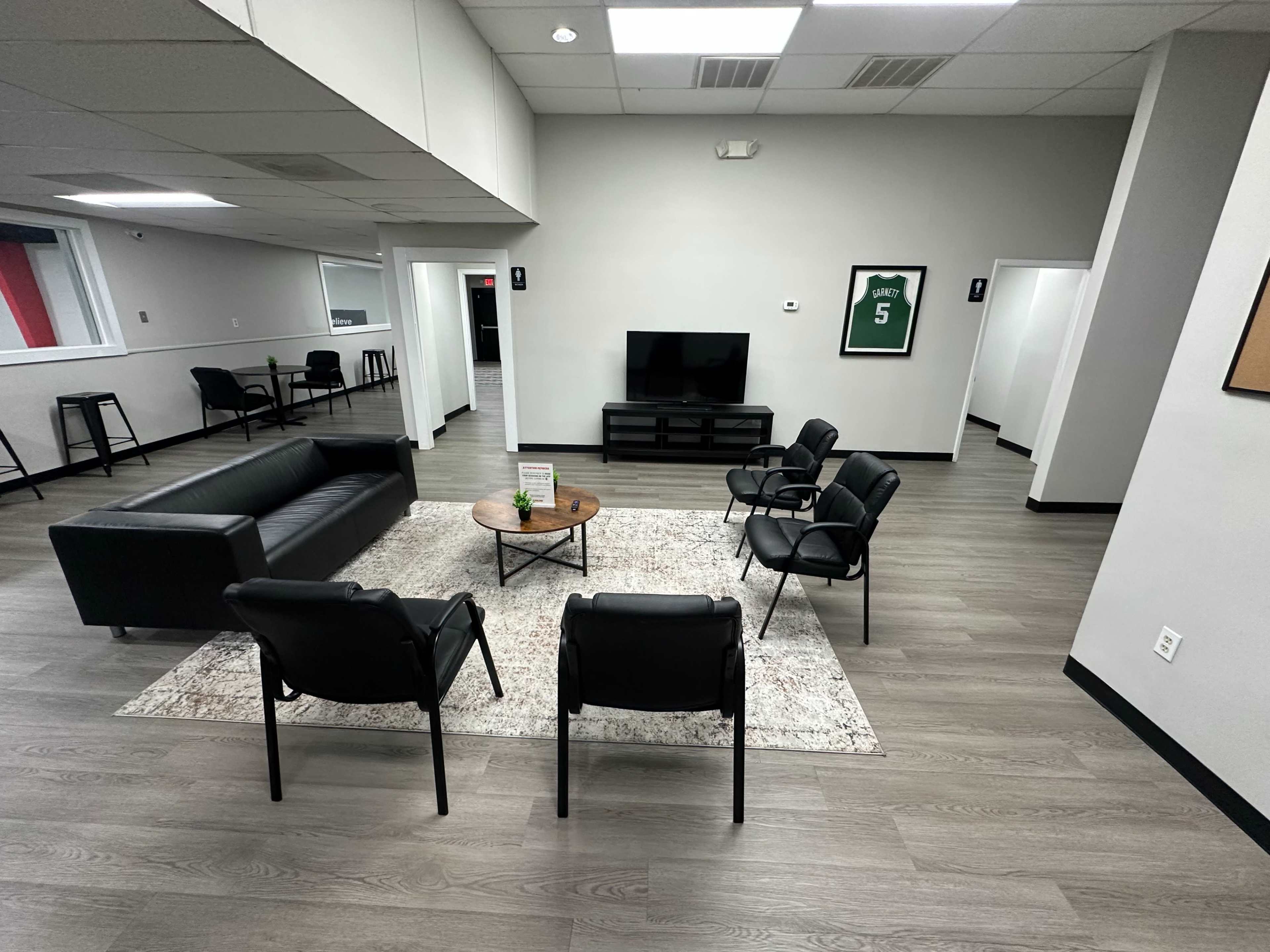 The image shows a modern, minimalist waiting area with a black sofa, four black chairs, a coffee table on a rug, and a television on a stand against a gray wall.