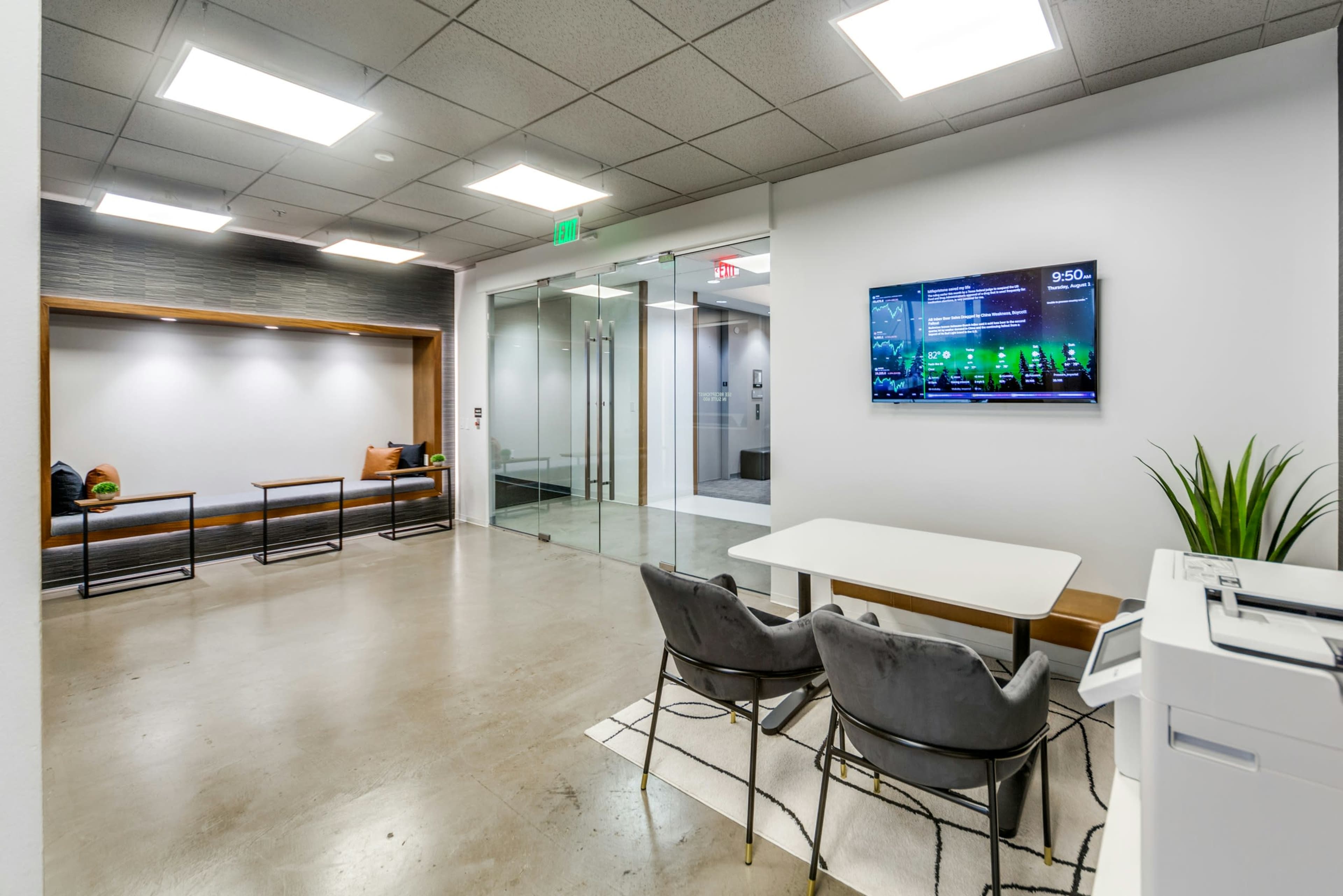 The image shows a modern office reception area with glass walls, a small table with chairs, and a wall-mounted display showing digital information.