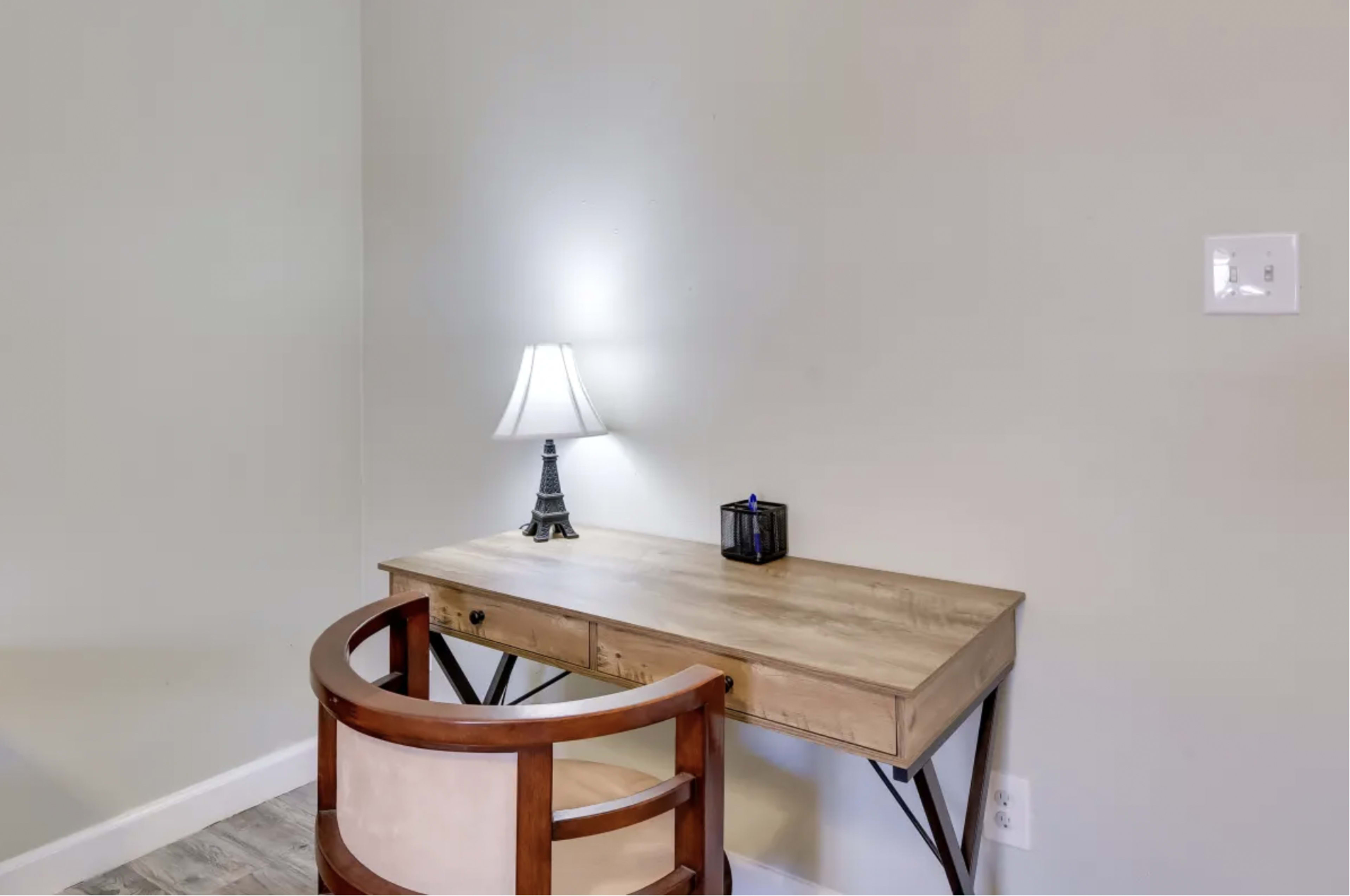 A wooden desk with a lamp and a chair is positioned against a plain wall.