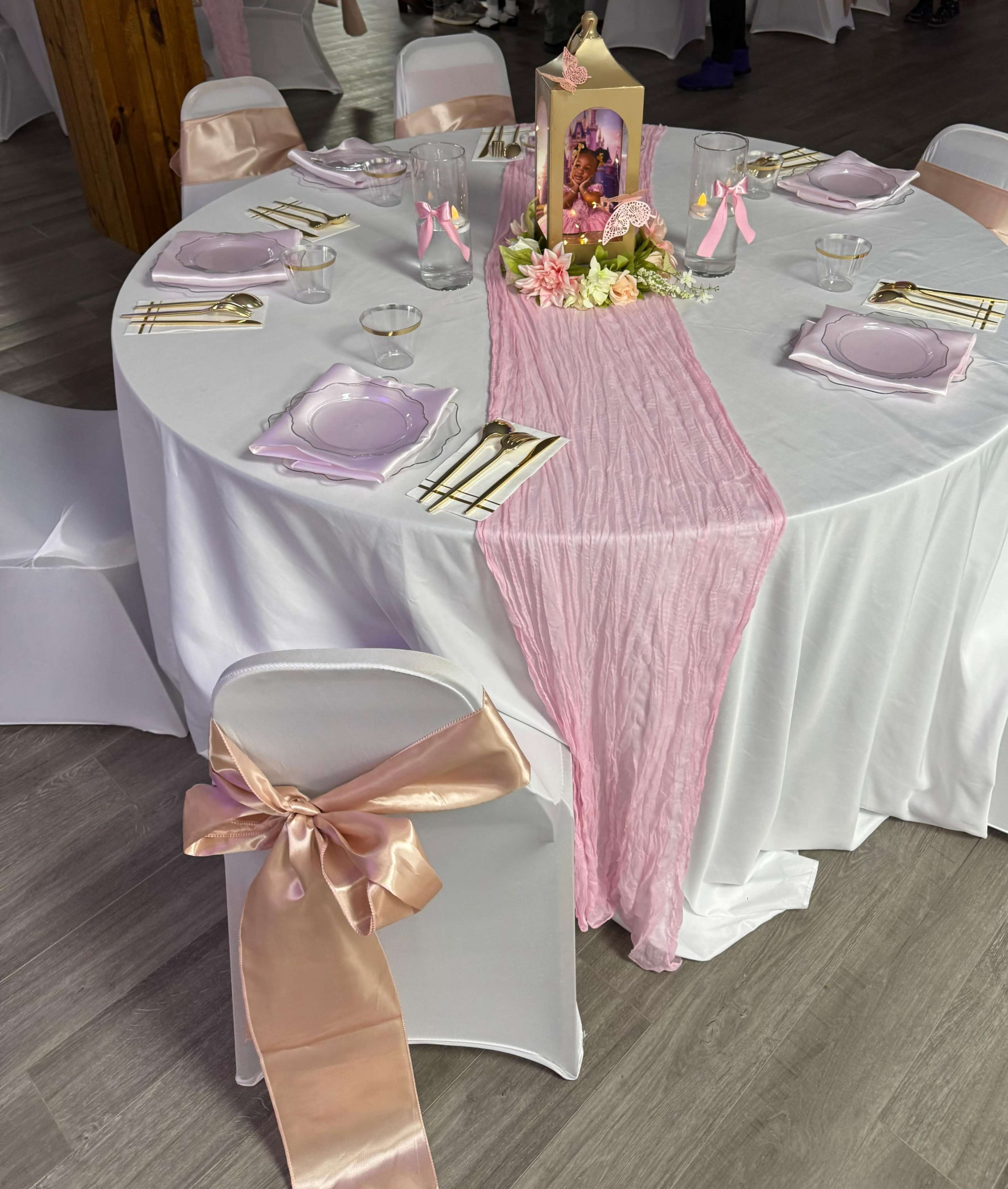 The image shows a round table covered with a white tablecloth, decorated with pink accents, plates, and a centerpiece featuring flowers and a framed photo.