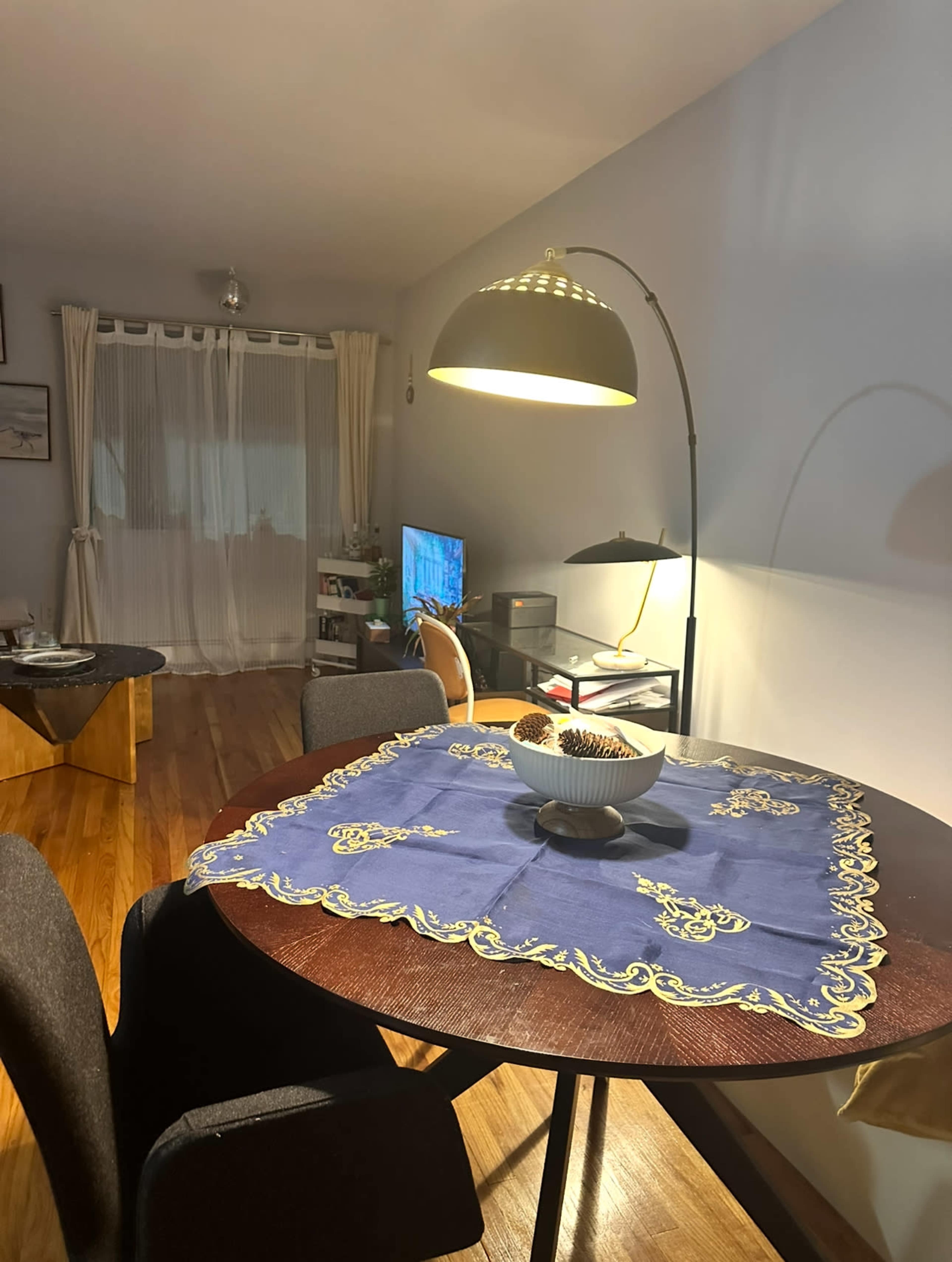 A dining table with a blue tablecloth, featuring a decorative bowl, is illuminated by a floor lamp, with a living space visible in the background.