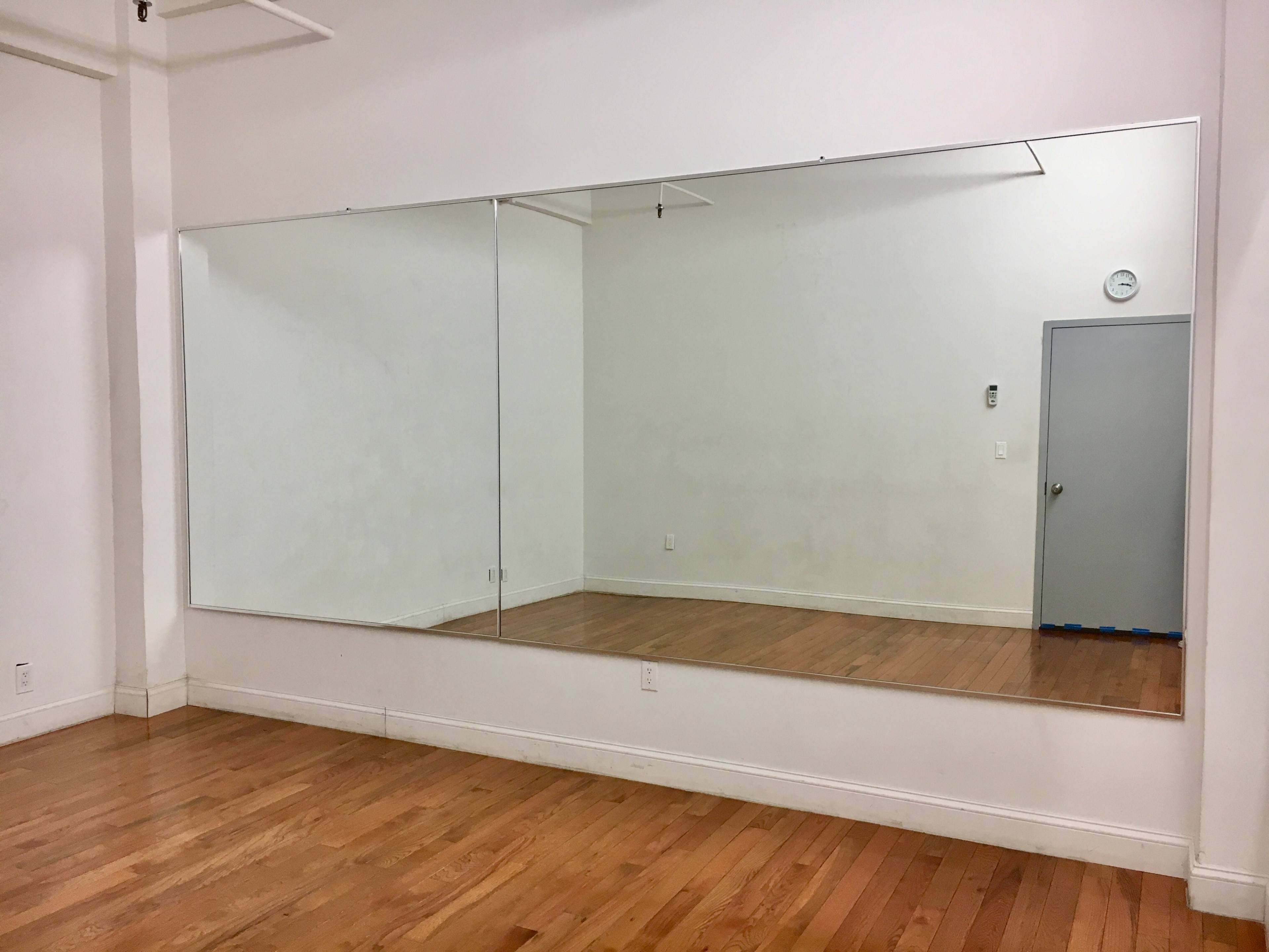 A spacious room features a large wall mirror above a wooden floor, with a door visible in the background.