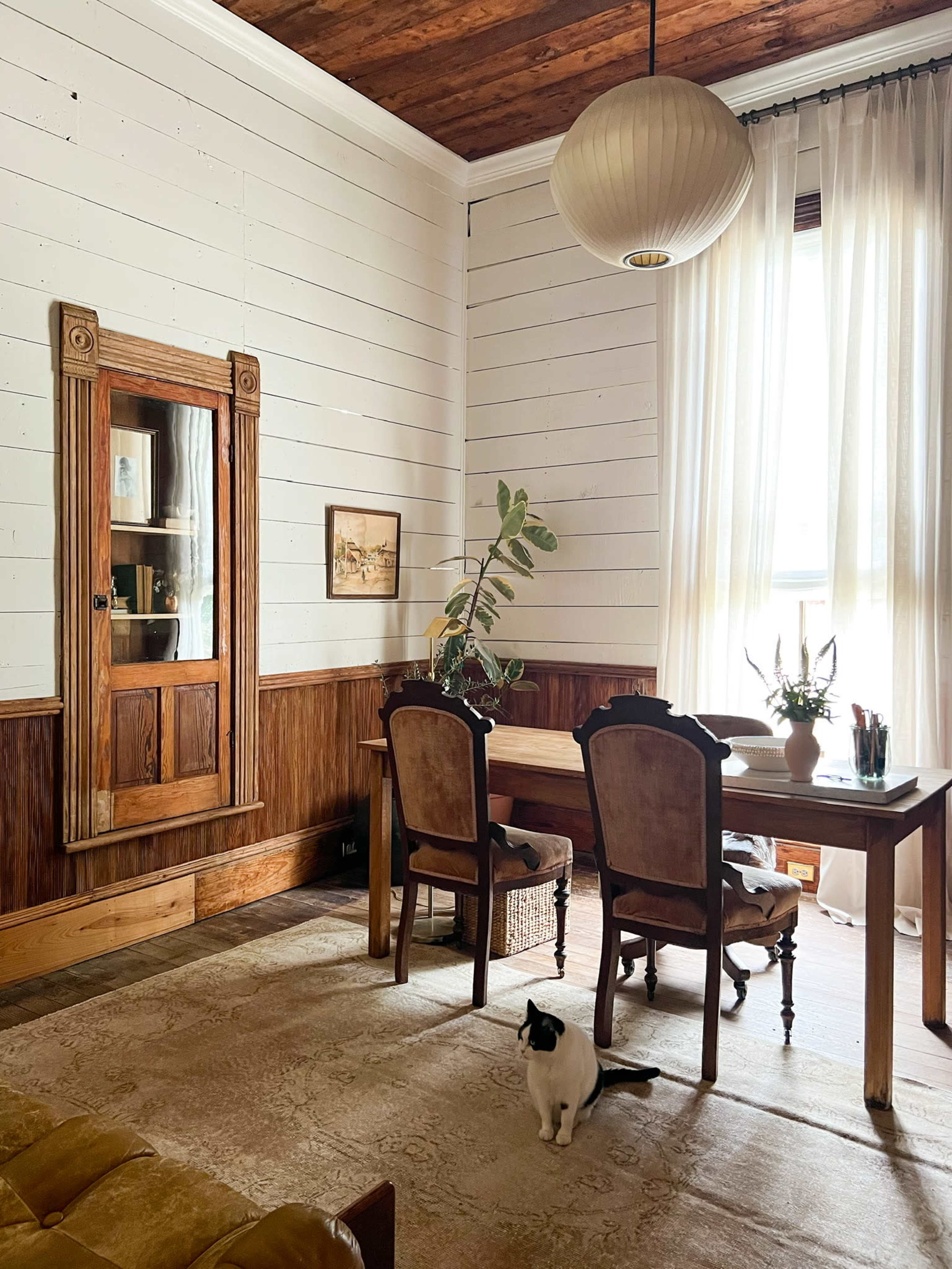 A spacious room features a dining table with two chairs, a large mirror with a wooden frame, and a cat on the floor, all illuminated by a pendant light and natural sunlight through sheer curtains.