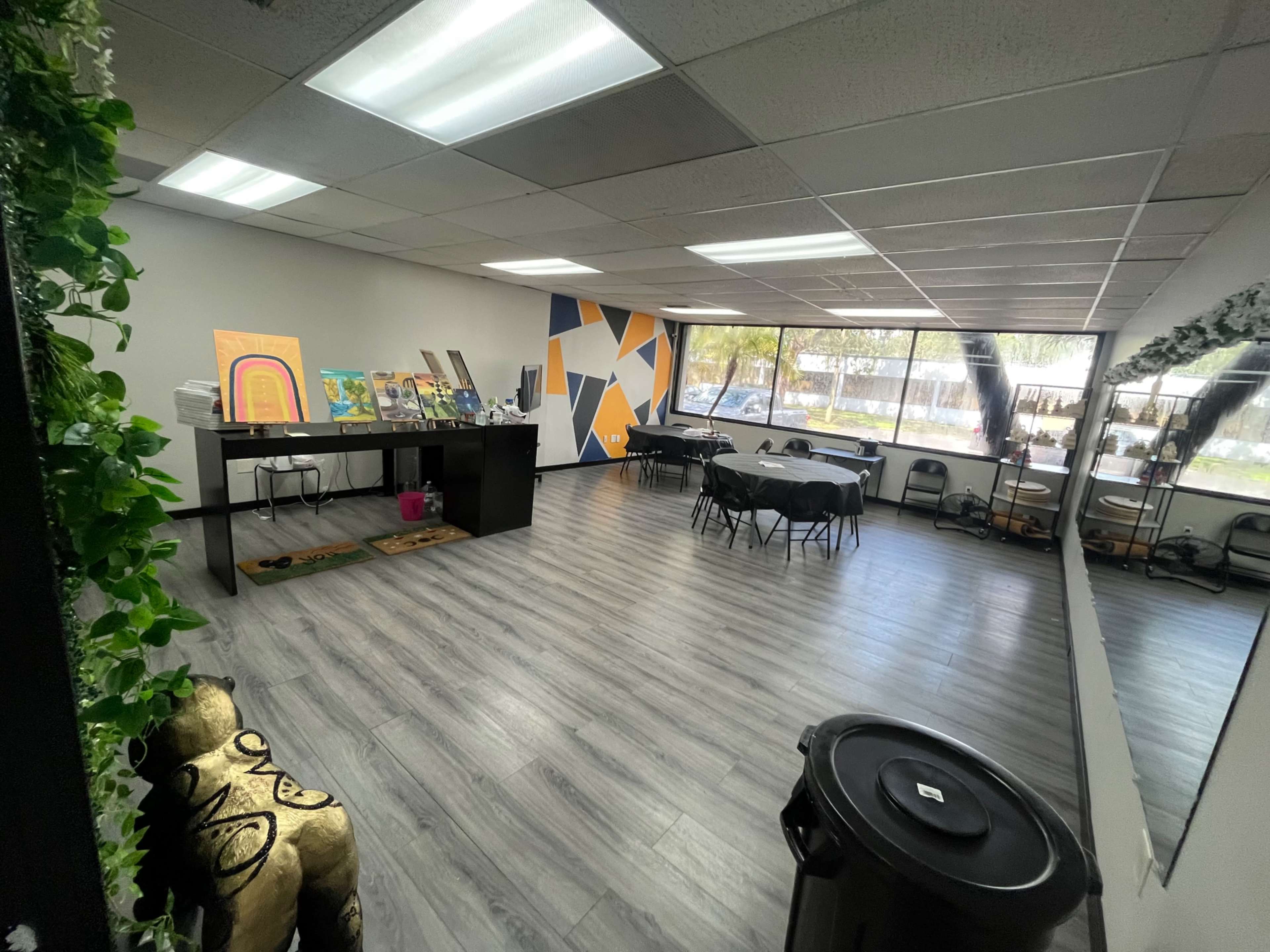 An empty office space with tables, chairs, artwork displayed on a counter, and large windows allowing natural light in.