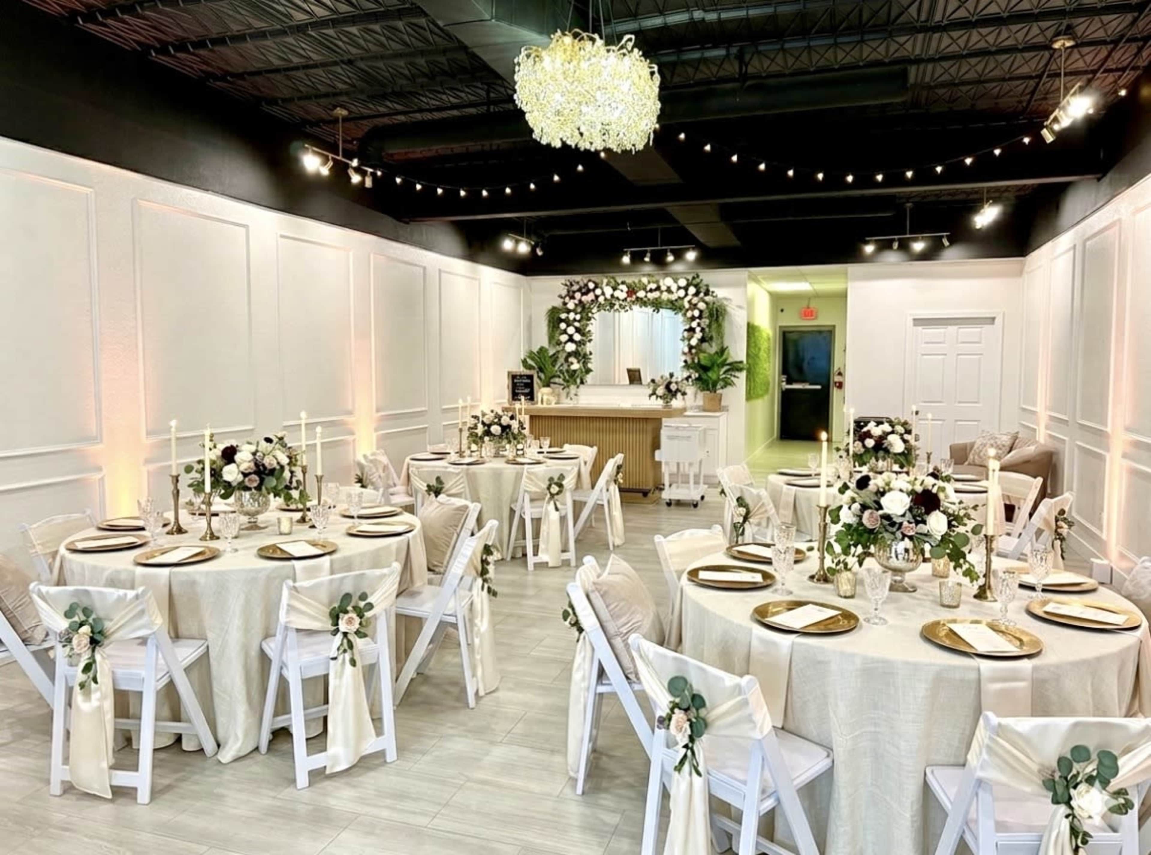 The image showcases a beautifully arranged event space featuring round dining tables with white chairs, adorned with floral centerpieces and elegant table settings, all under soft lighting and decorative elements.