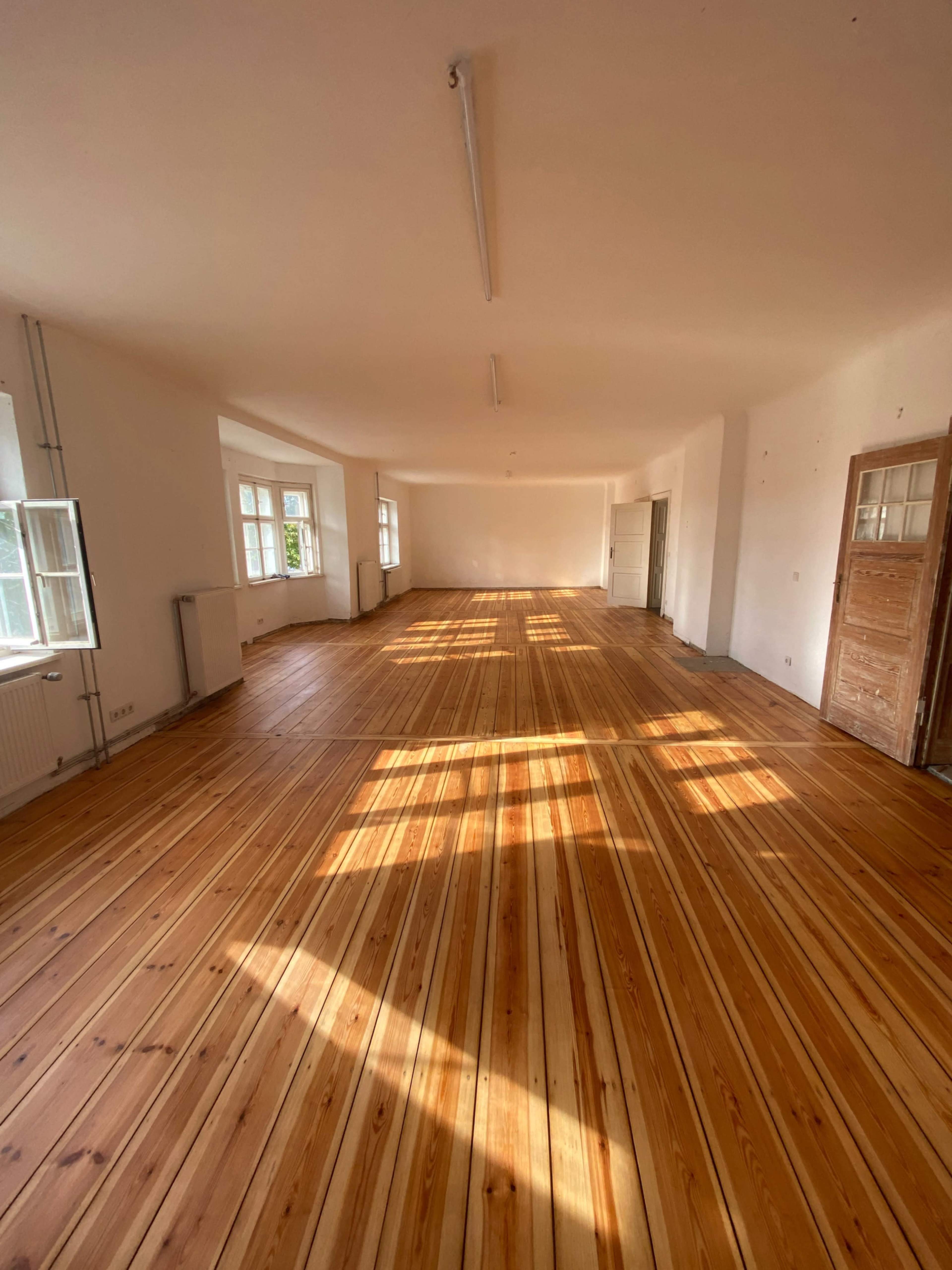 The image shows a spacious room with wooden flooring and large windows allowing natural light to fill the space.