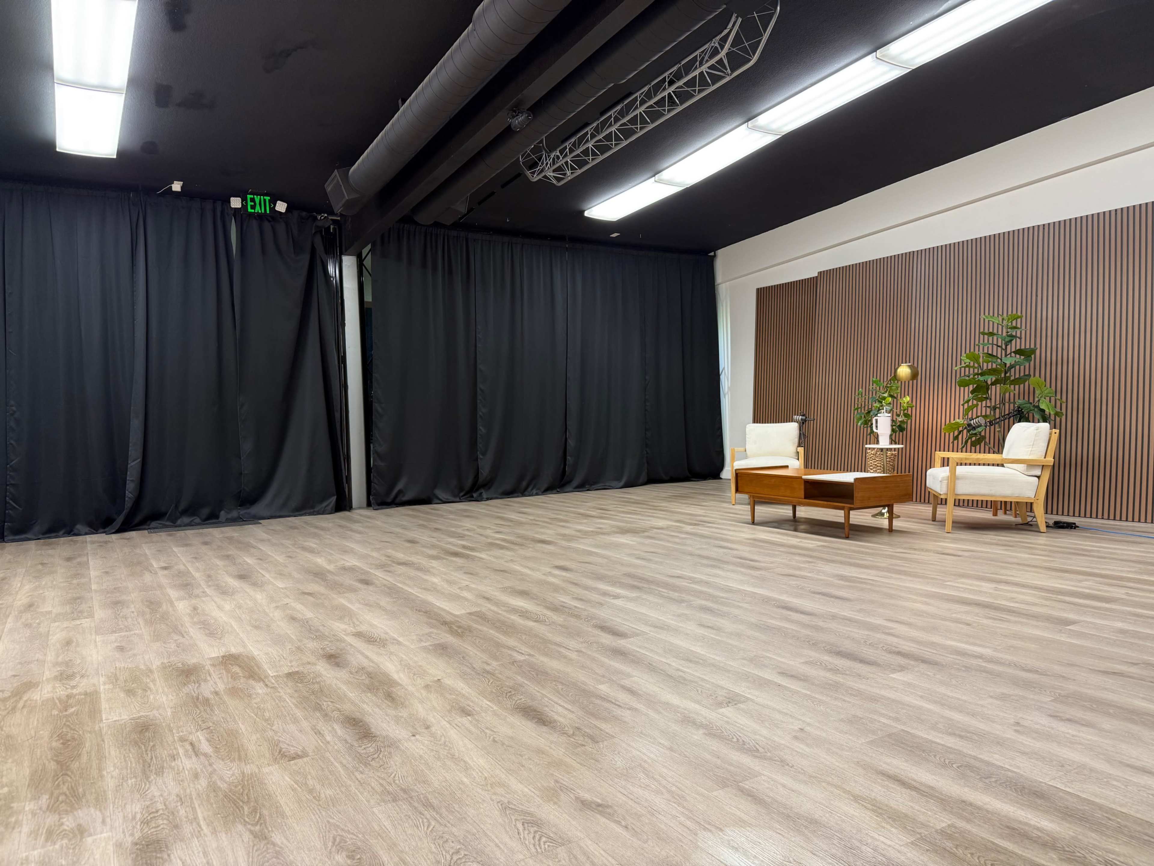 The image shows a spacious, empty room with black curtains on one side, hardwood flooring, and a small seating area consisting of two chairs and a coffee table.