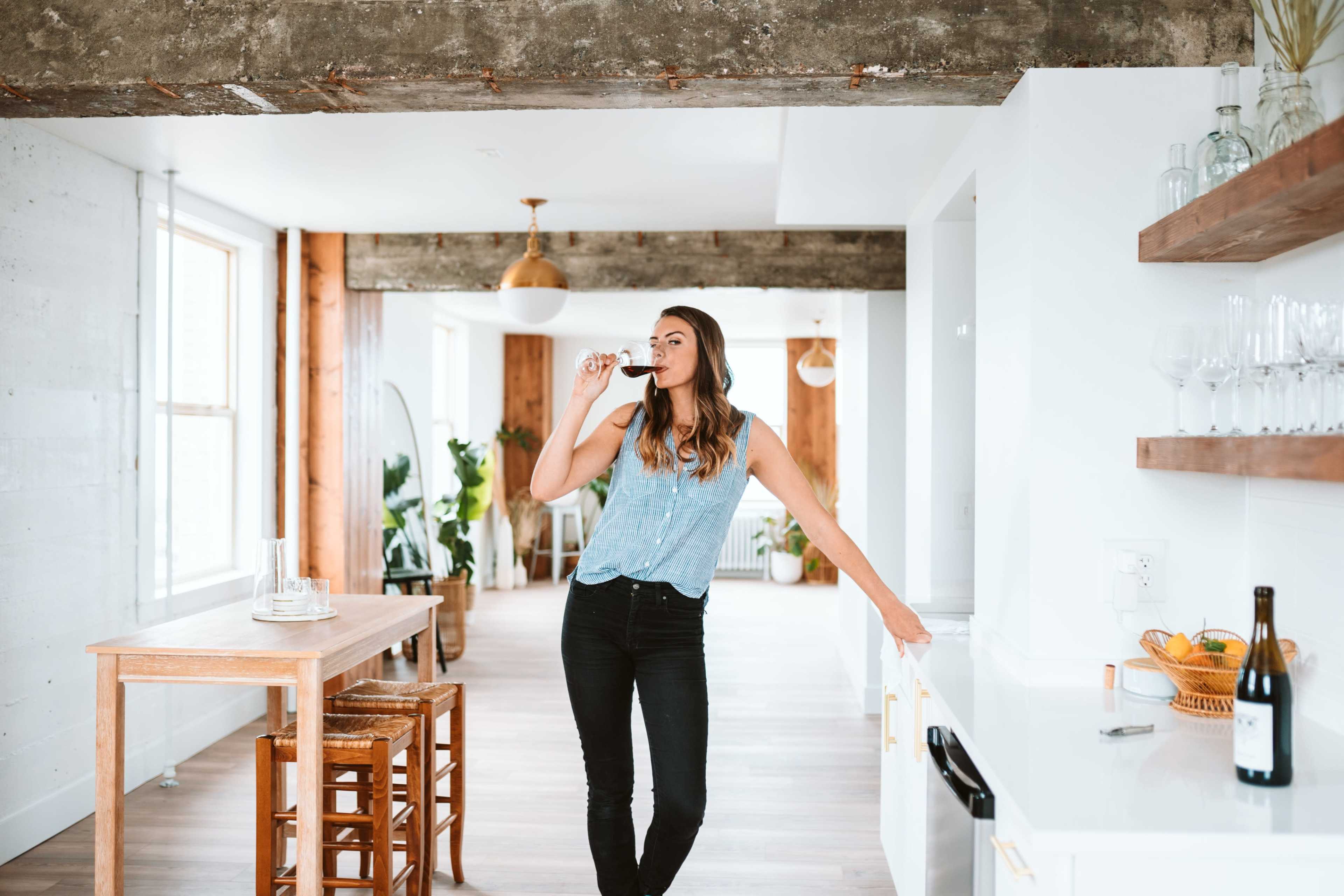 A woman stands in a modern kitchen, sipping from a wine glass while leaning against a white countertop.