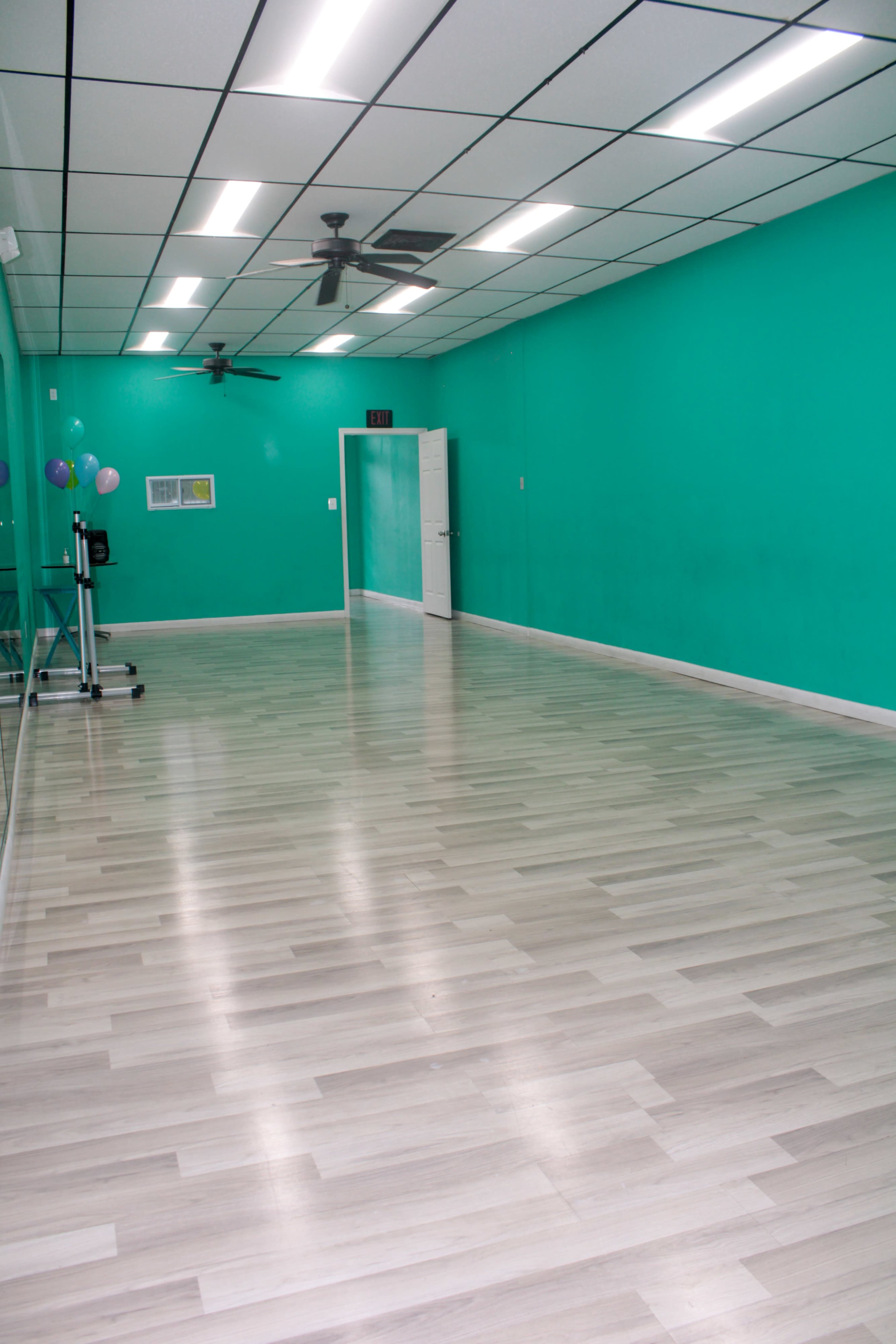 A large, empty room with a teal wall, white ceiling tiles, and light wooden flooring.