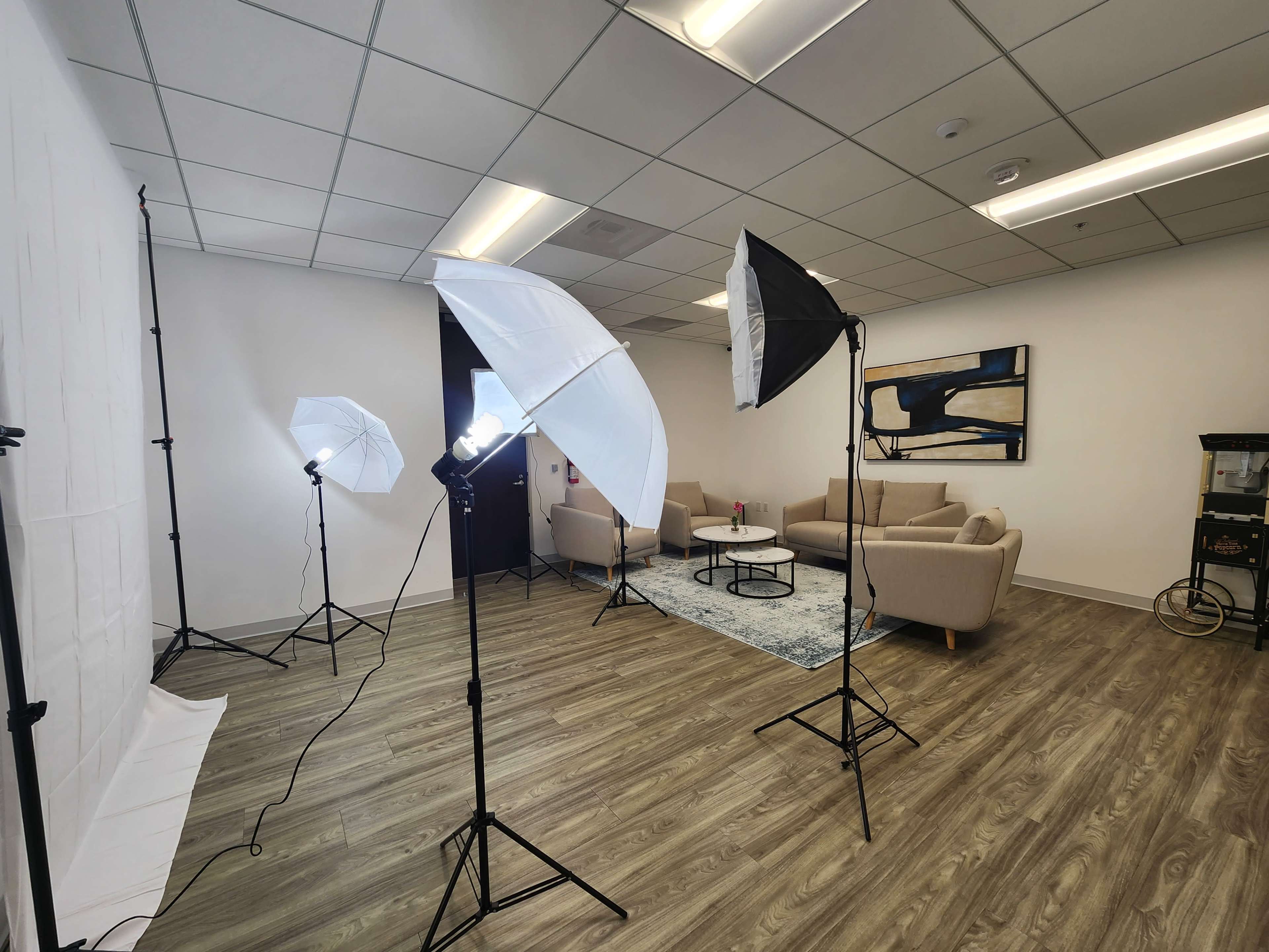 A well-lit photo studio setup with two light umbrellas and two softboxes positioned around a seating area featuring two beige sofas, a coffee table, and a decorative rug.
