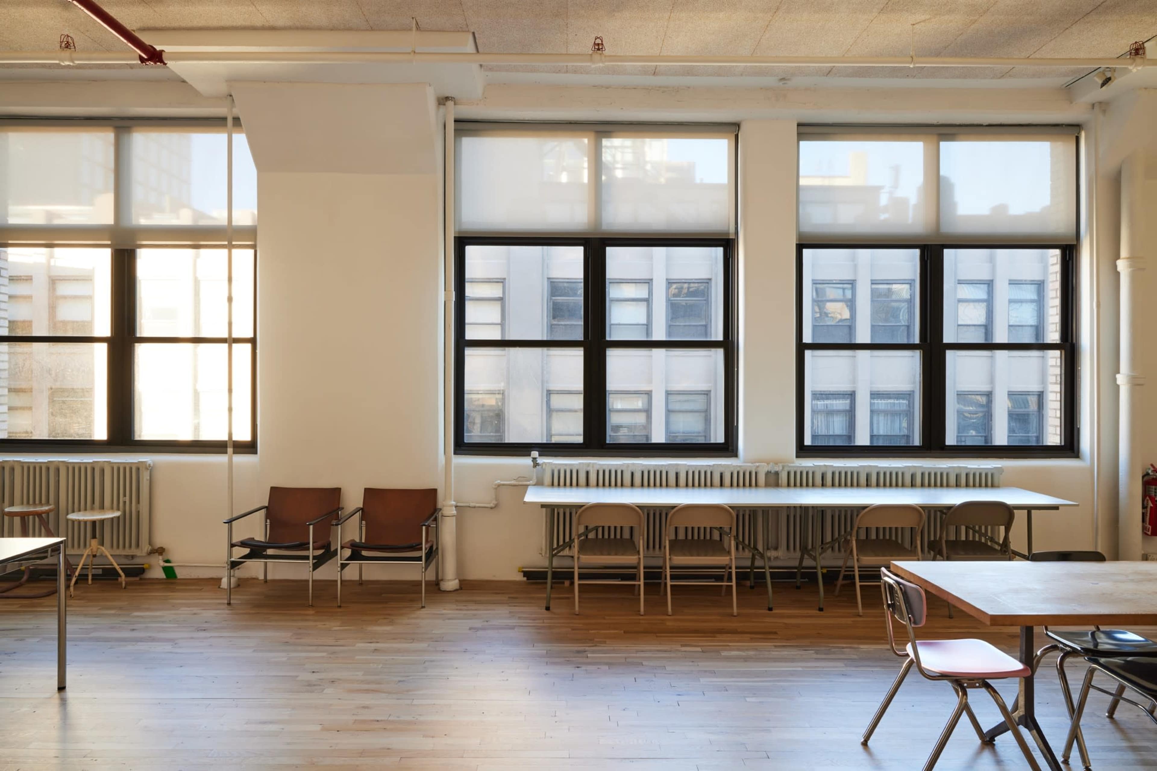 The image shows a spacious room with large windows, wooden floors, and a row of chairs and tables along the walls.