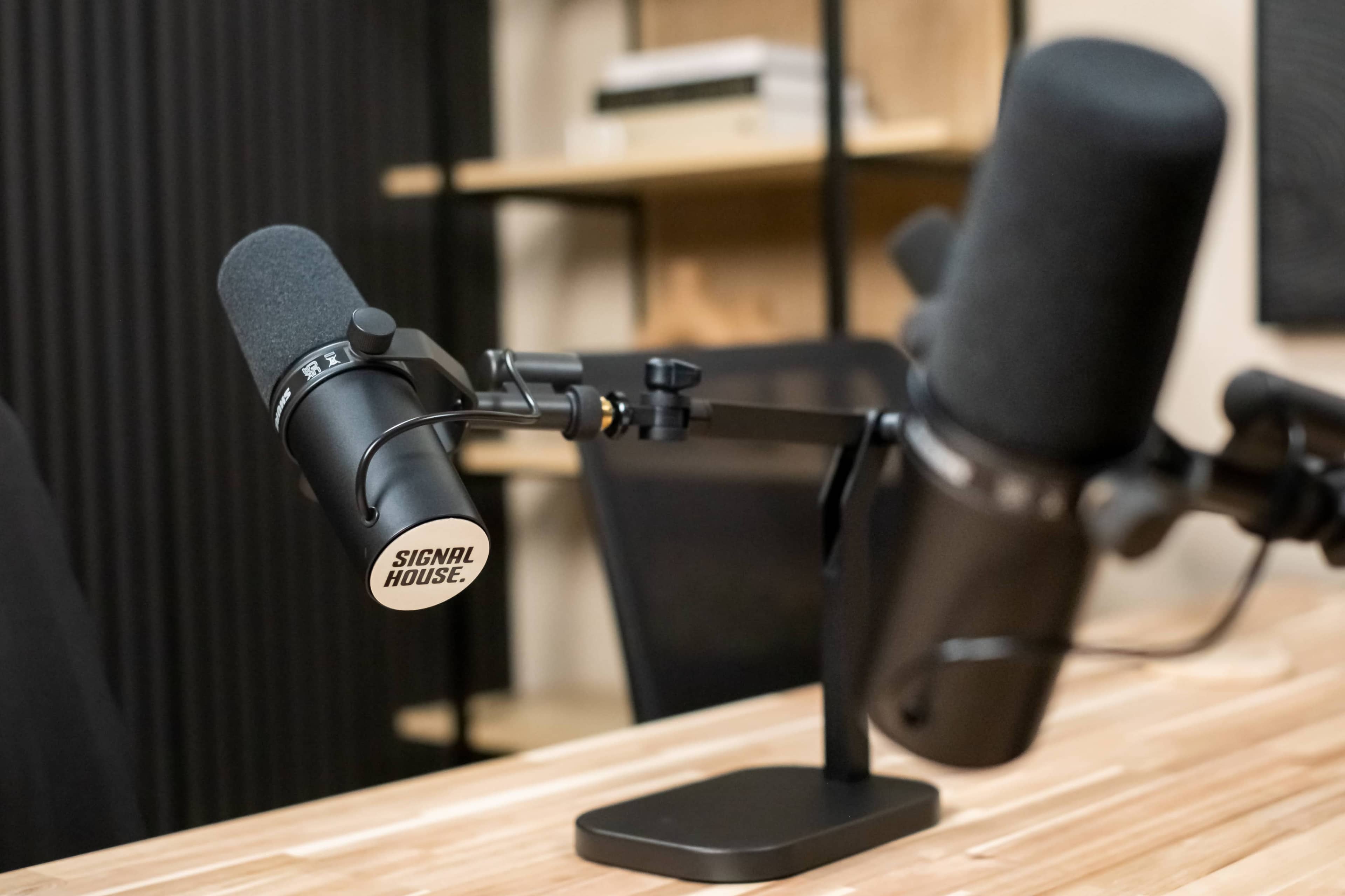 The image shows two microphones mounted on stands, positioned on a wooden table in a room with a dark background and shelving.
