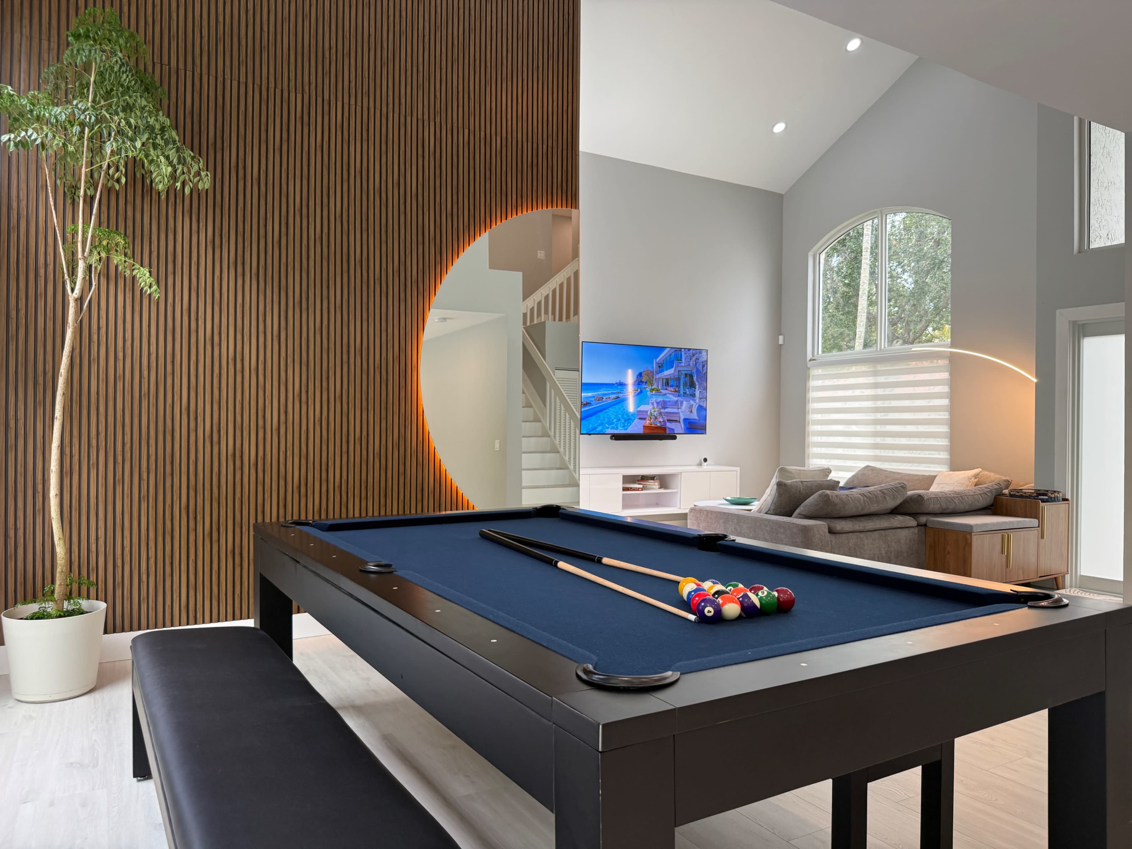 The image features a modern interior with a billiard table in the foreground, a wall with vertical wood paneling, a large television mounted on a light-colored wall, and a staircase in the background.