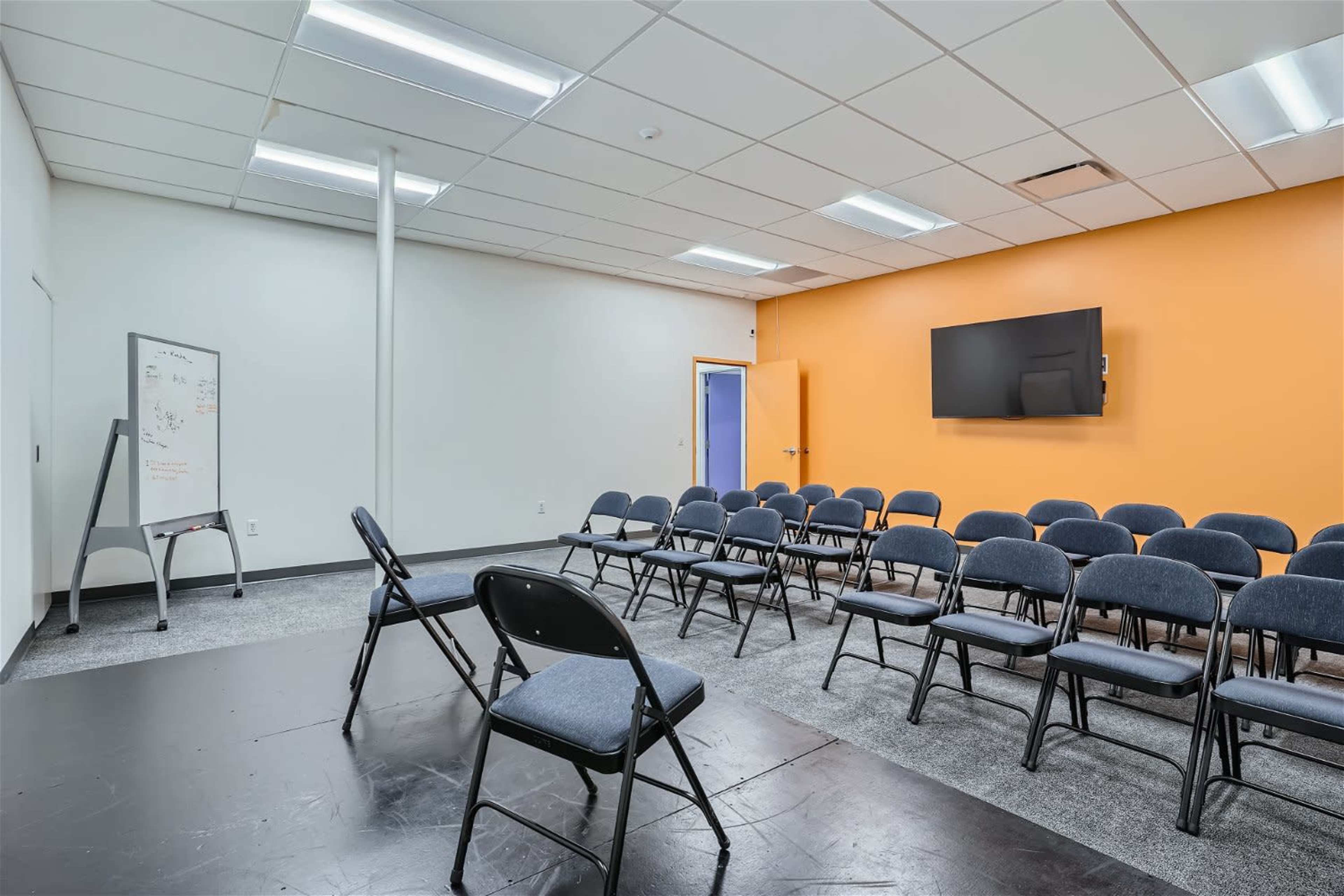 A meeting room with rows of black chairs, a whiteboard, and a wall-mounted television, featuring an orange accent wall.