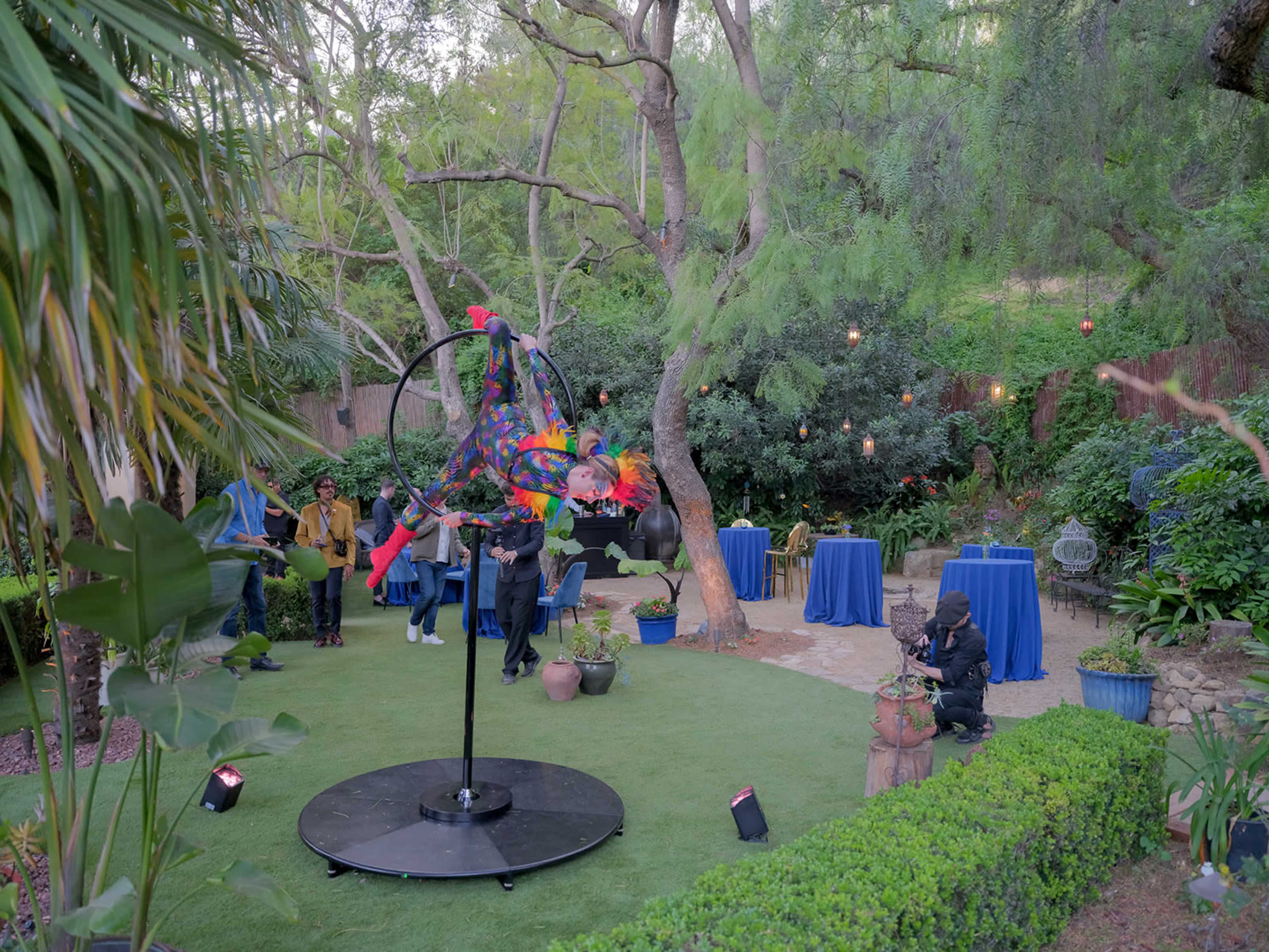 An outdoor gathering features a colorful performer suspended in mid-air on a hoop, surrounded by guests and decorated tables in a lush garden setting.