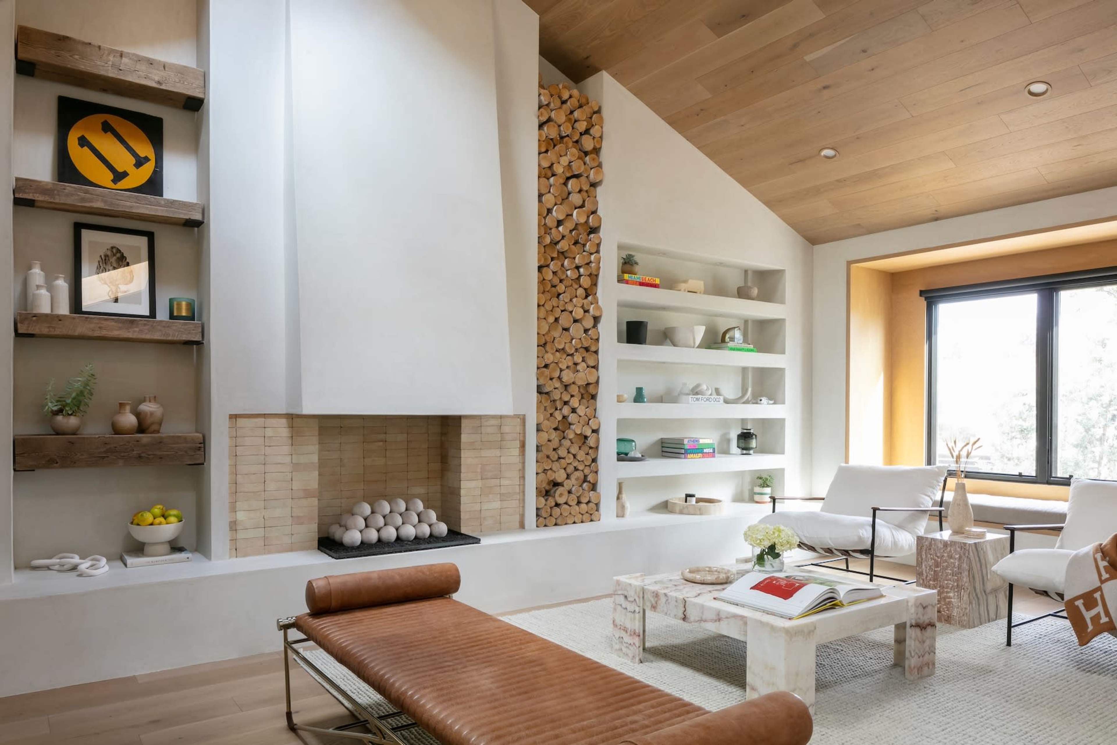 A modern living room featuring a fireplace with stacked wooden logs, a light-colored wall, and large windows letting in natural light.