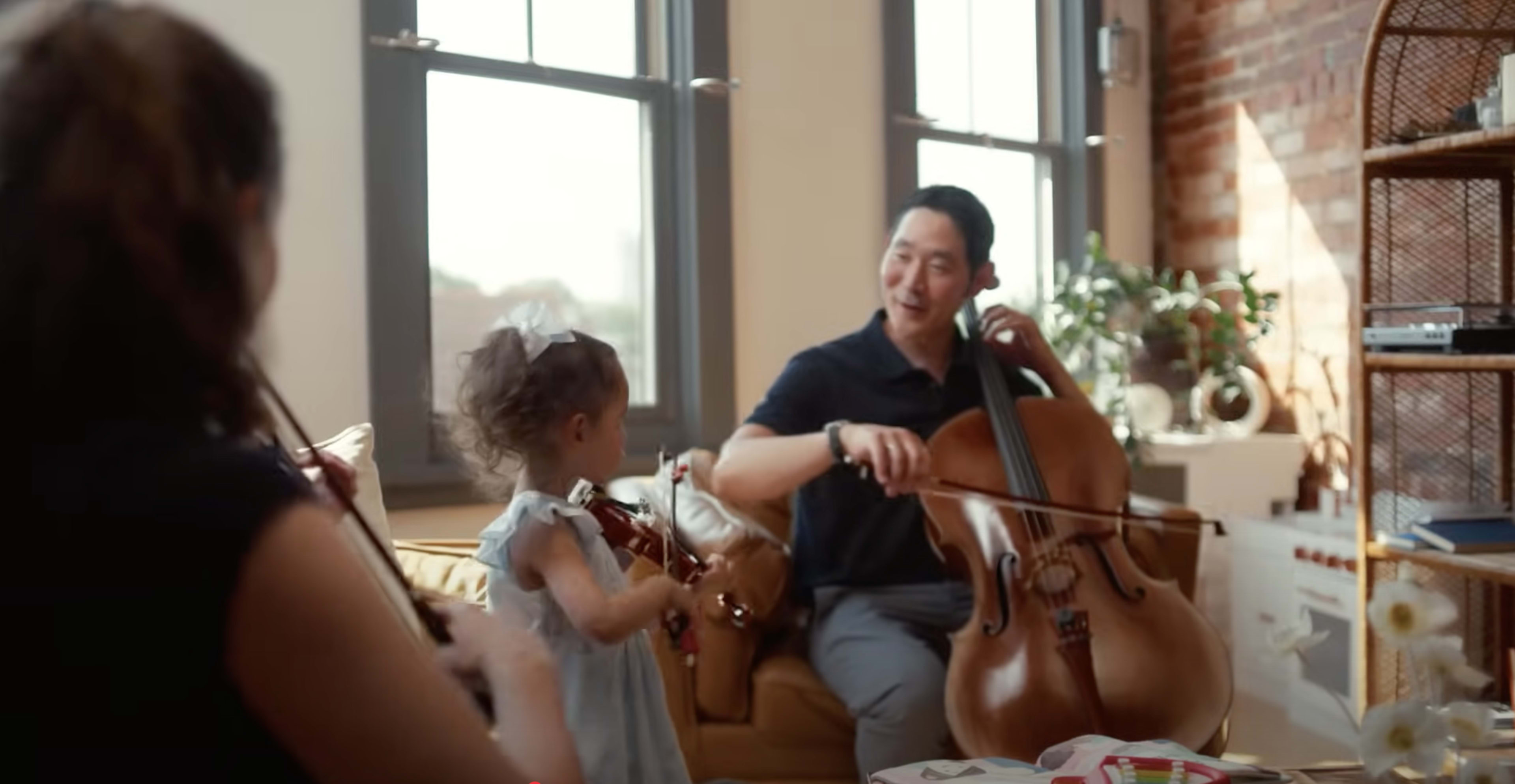 A man plays the cello while a young girl holding a violin smiles at him in a sunlit living room.