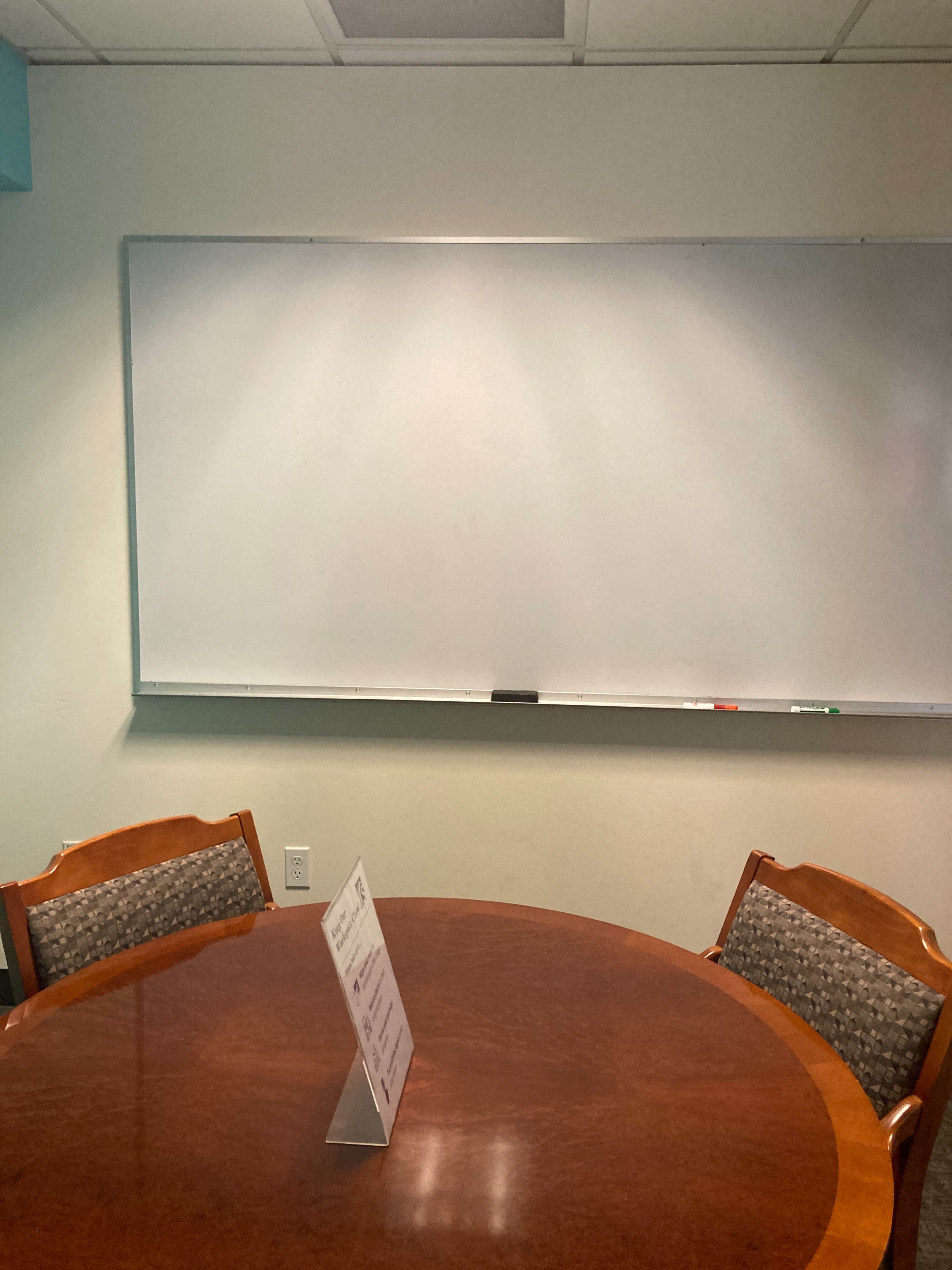 A round wooden table with two chairs sits in front of a blank whiteboard in a meeting room.
