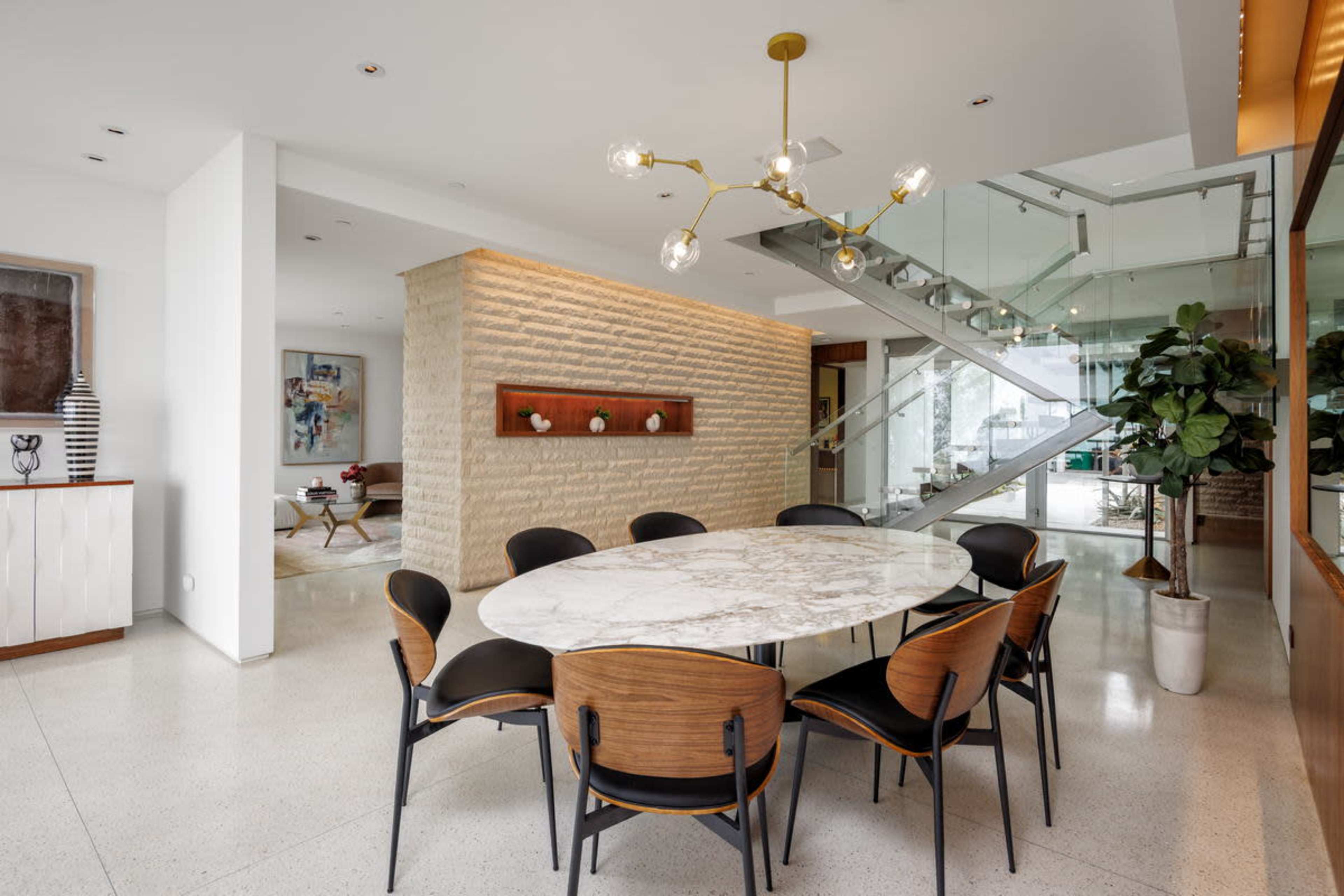 A modern dining area features a round marble table surrounded by black and wooden chairs, with a staircase and a textured stone wall in the background.