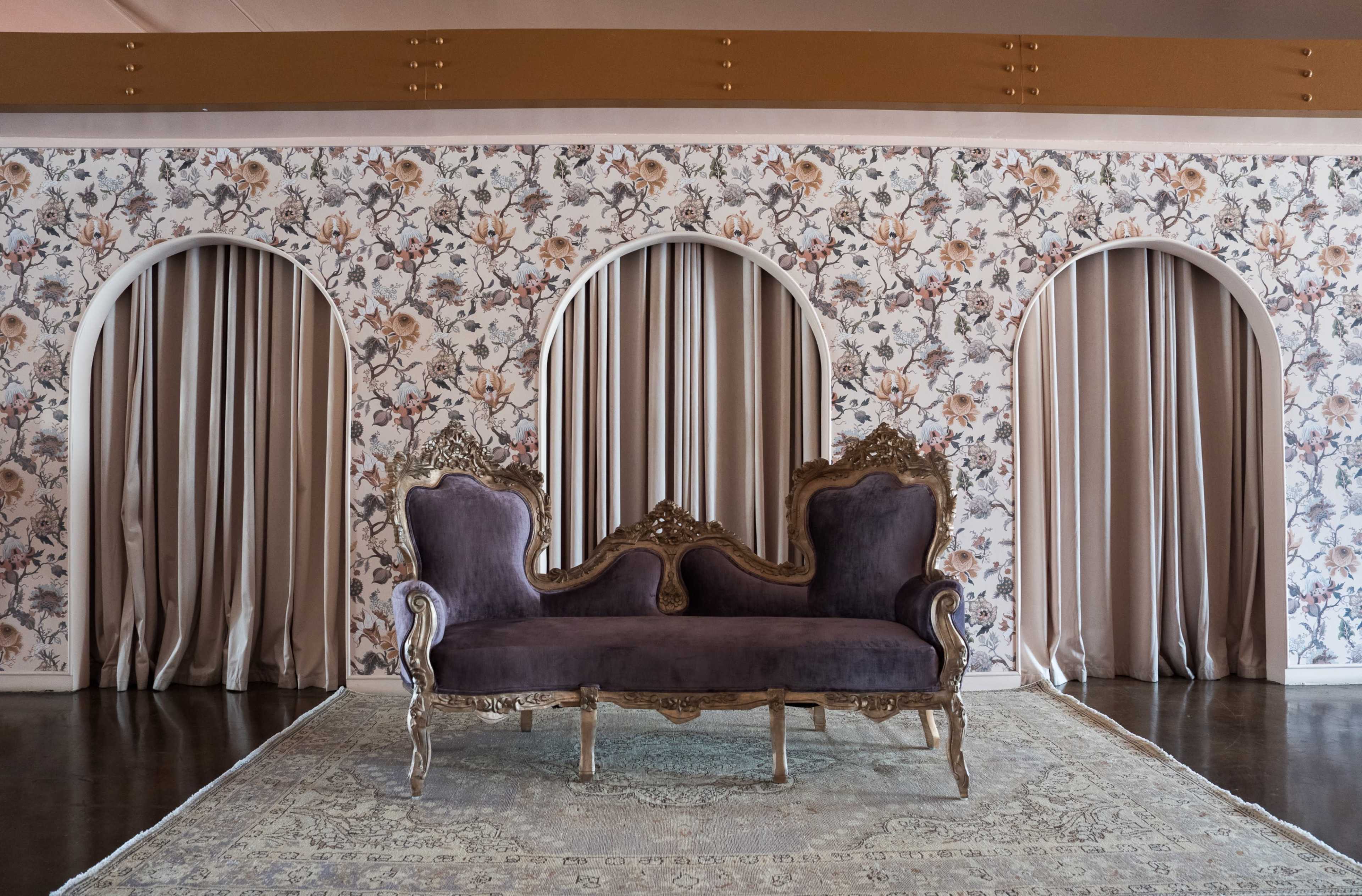 A vintage purple sofa is placed in front of a floral-patterned wall featuring three arched alcoves with fabric drapery.