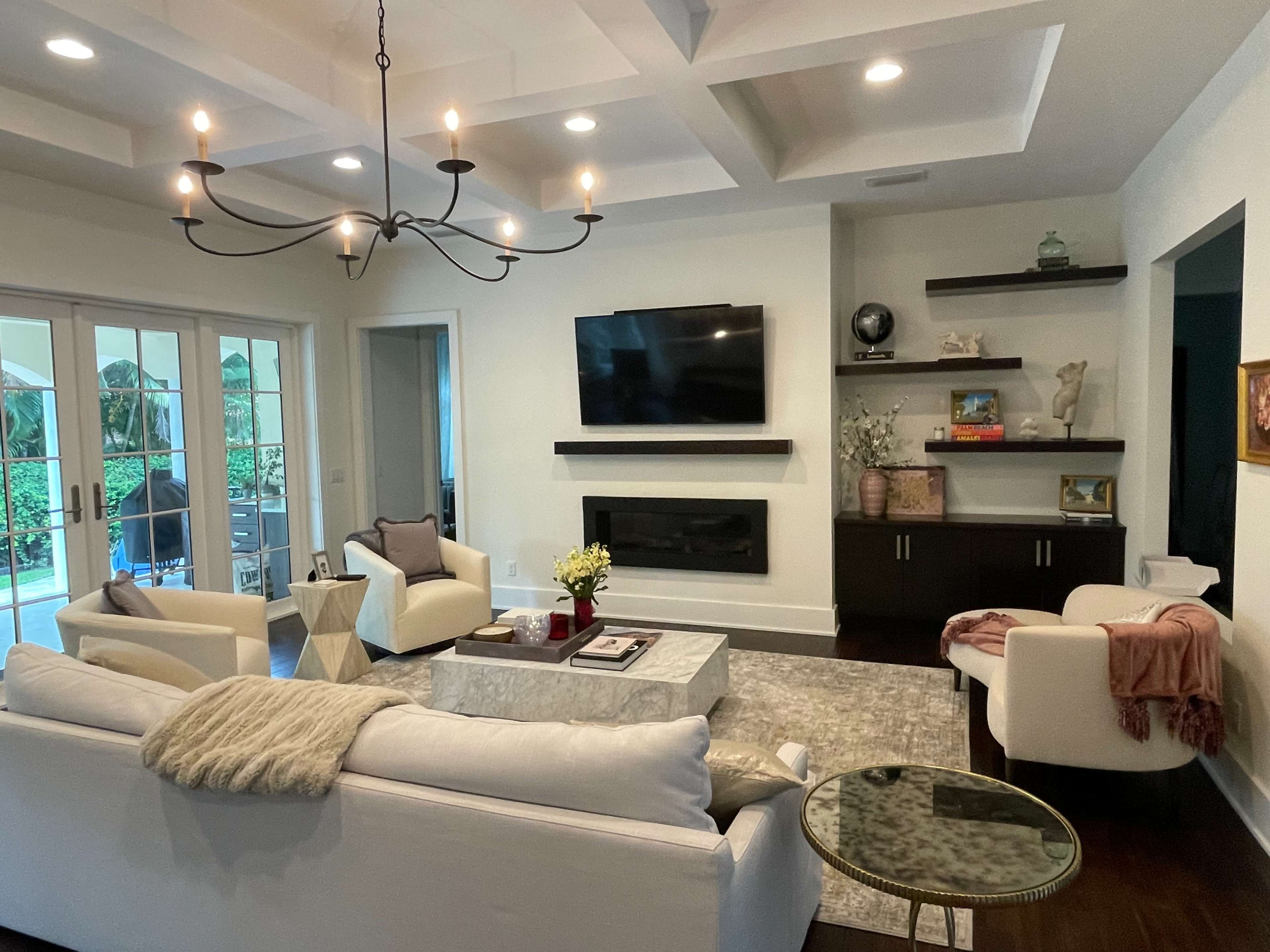 A modern living room with a white sofa, a coffee table, a television mounted on the wall, and a chandelier, all arranged around a space that features large windows and a view of greenery outside.
