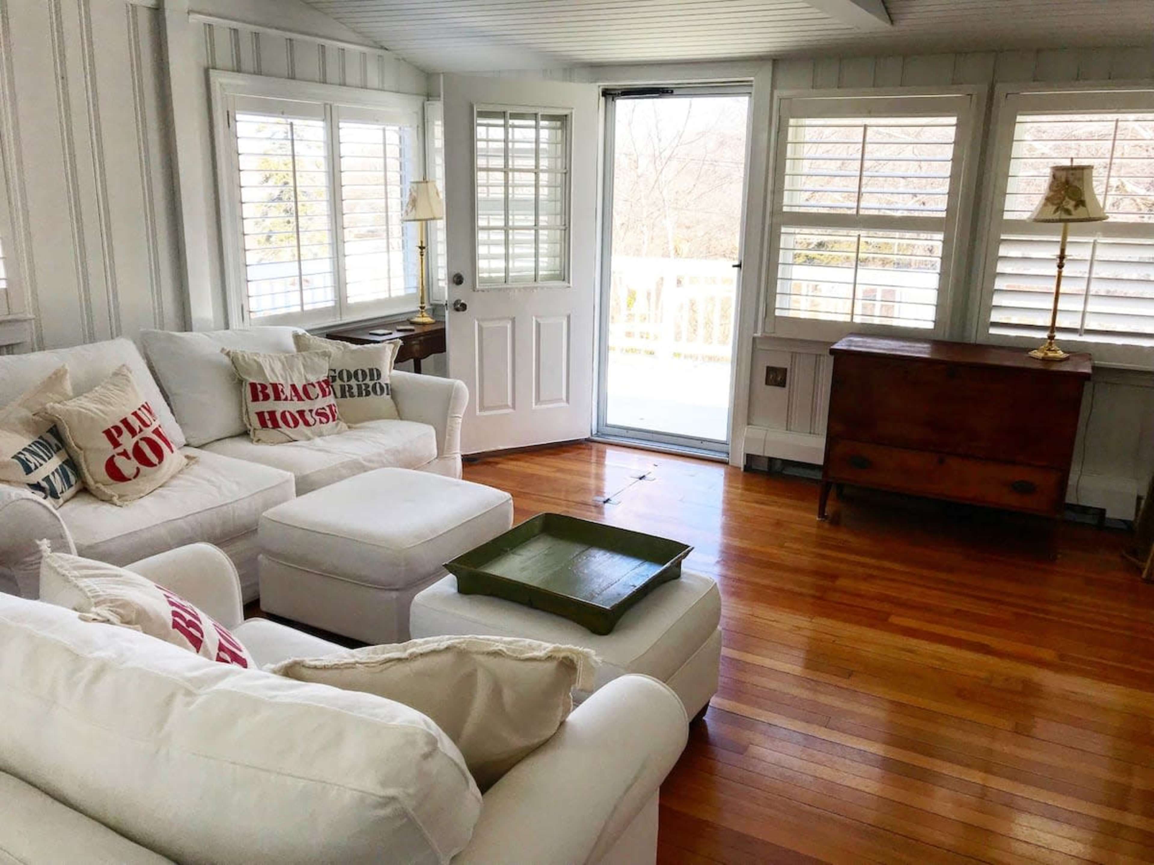 A bright, airy living room with a white sofa, decorative cushions, a wooden chest, and large windows that open to a balcony.