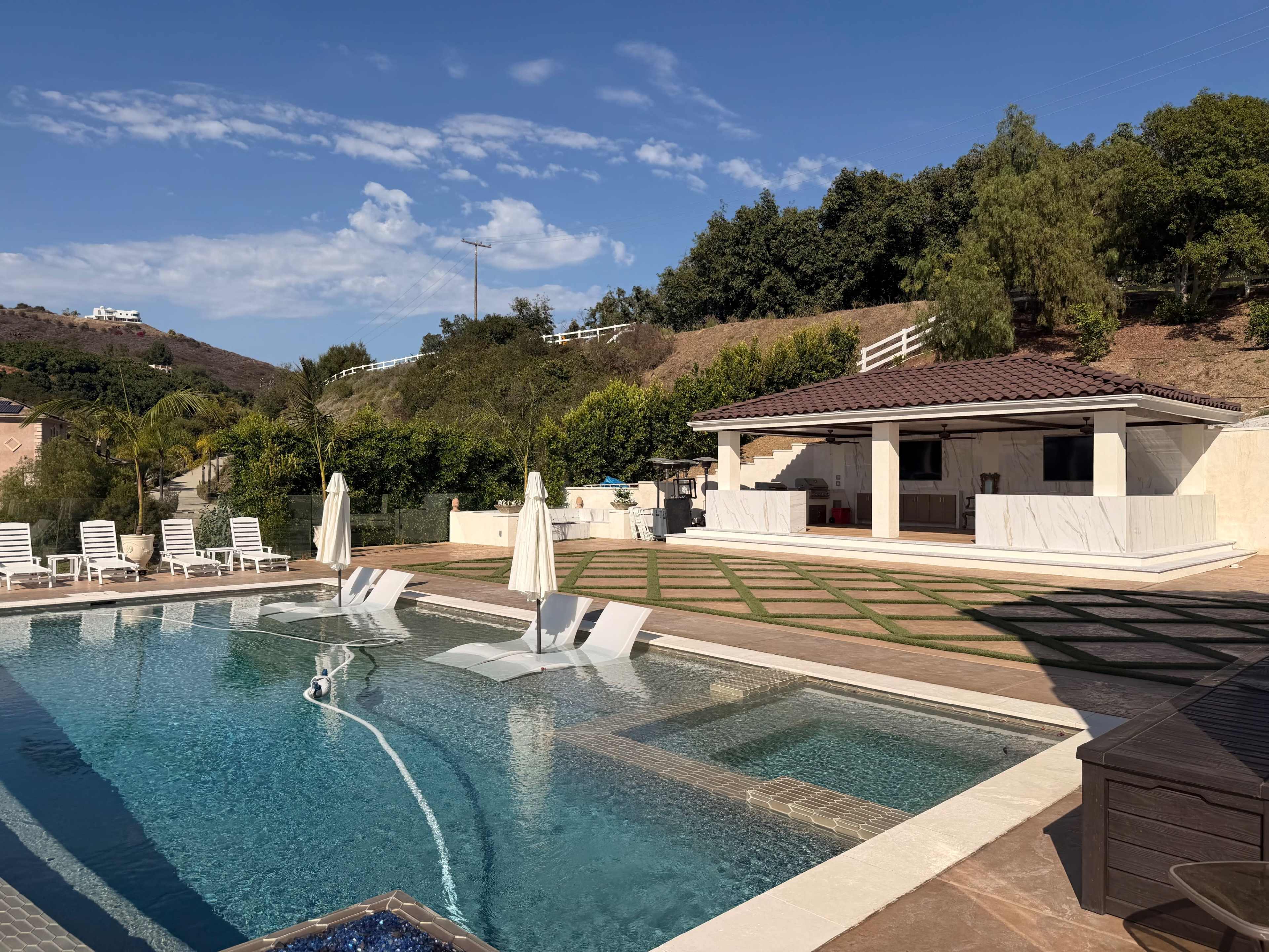 The image shows a large swimming pool surrounded by lounge chairs and a patio area with a covered structure in the background, set against a hillside landscape.