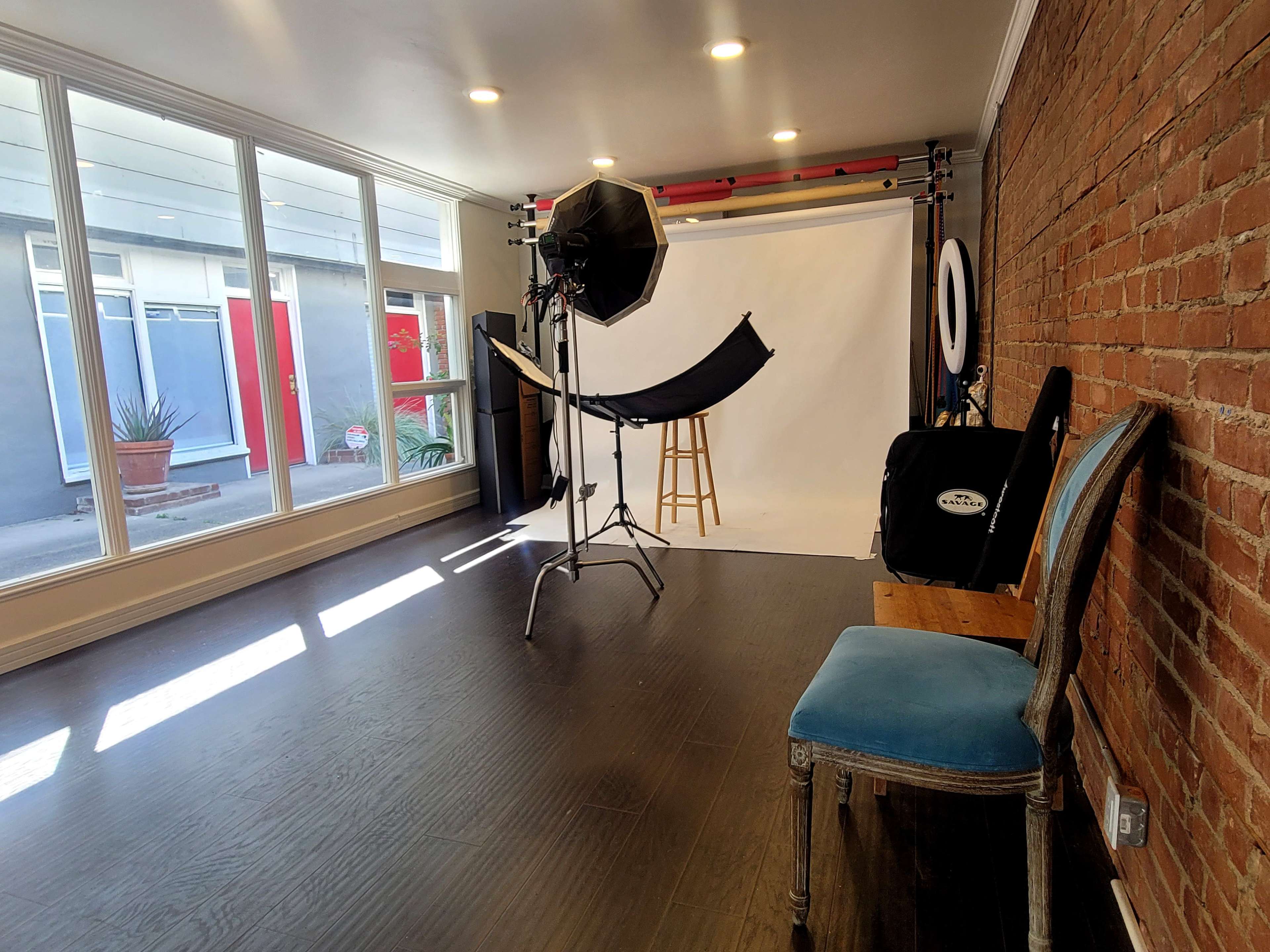 The image shows a photography studio with a backdrop, a stool, a lighting setup, and a chair against a brick wall.