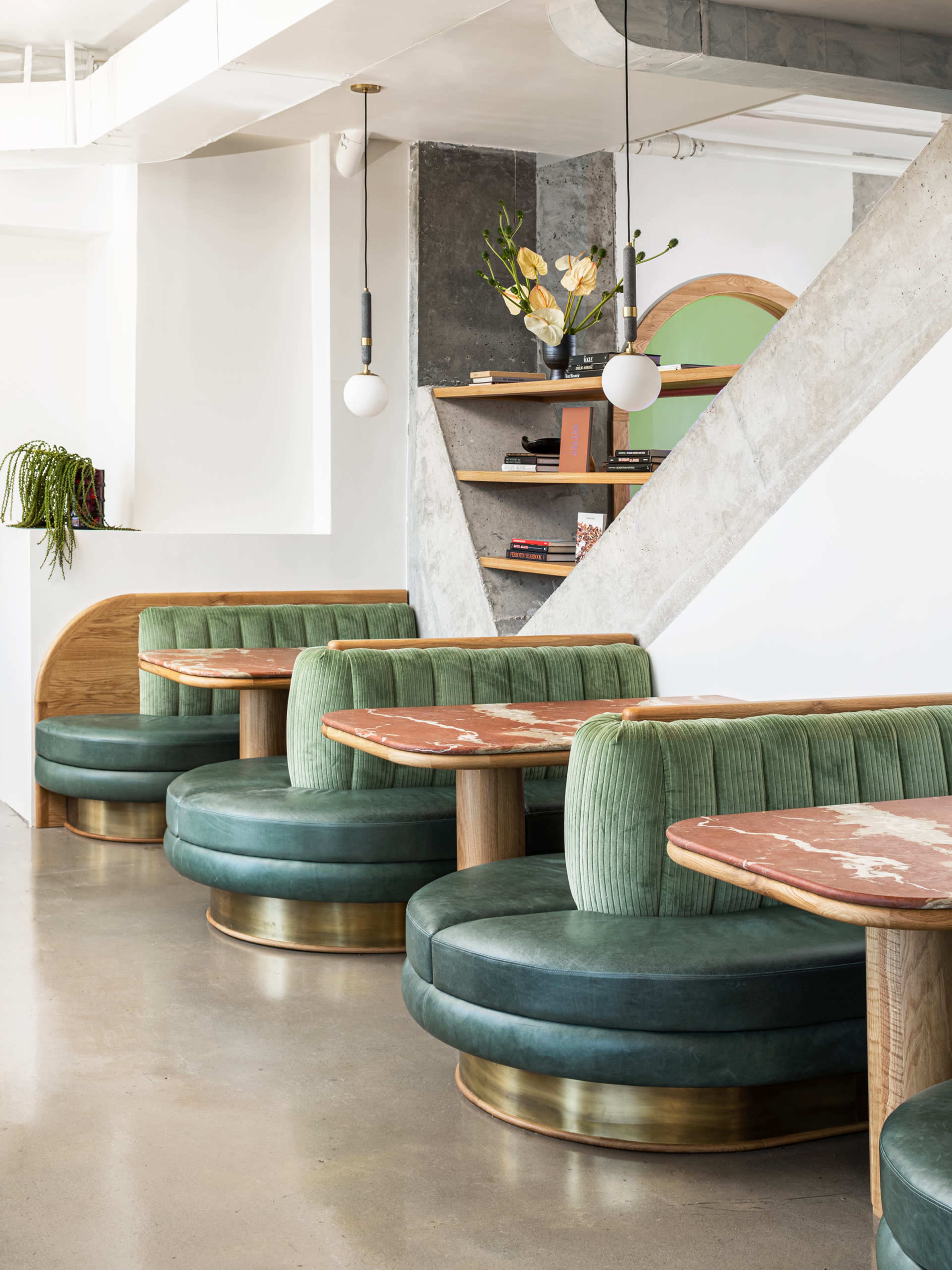 A modern dining area with green velvet booths and round wooden tables, complemented by a minimalist decor and large windows.