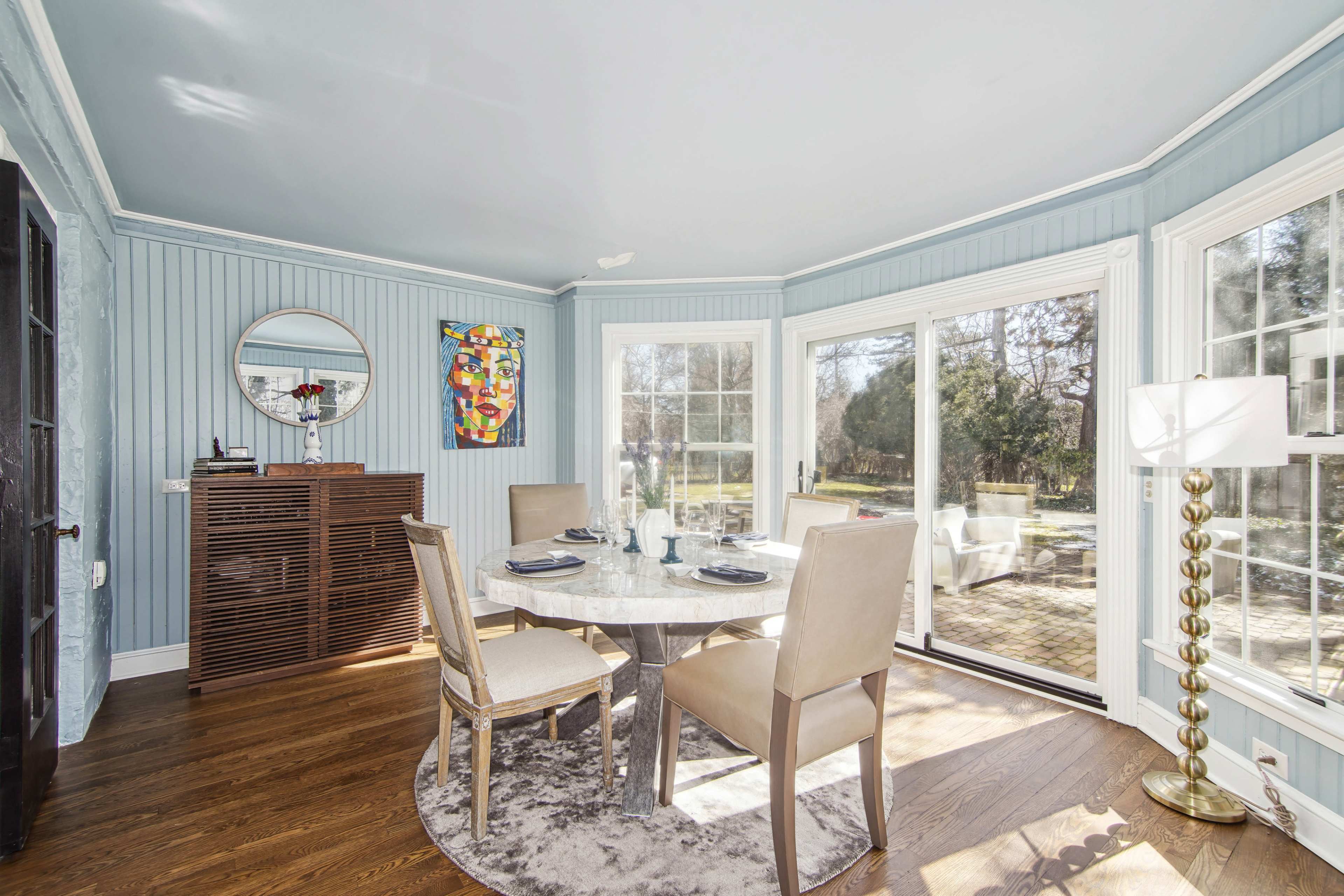 A bright dining area featuring a round marble table with four chairs, a side cabinet, and large windows overlooking a garden.