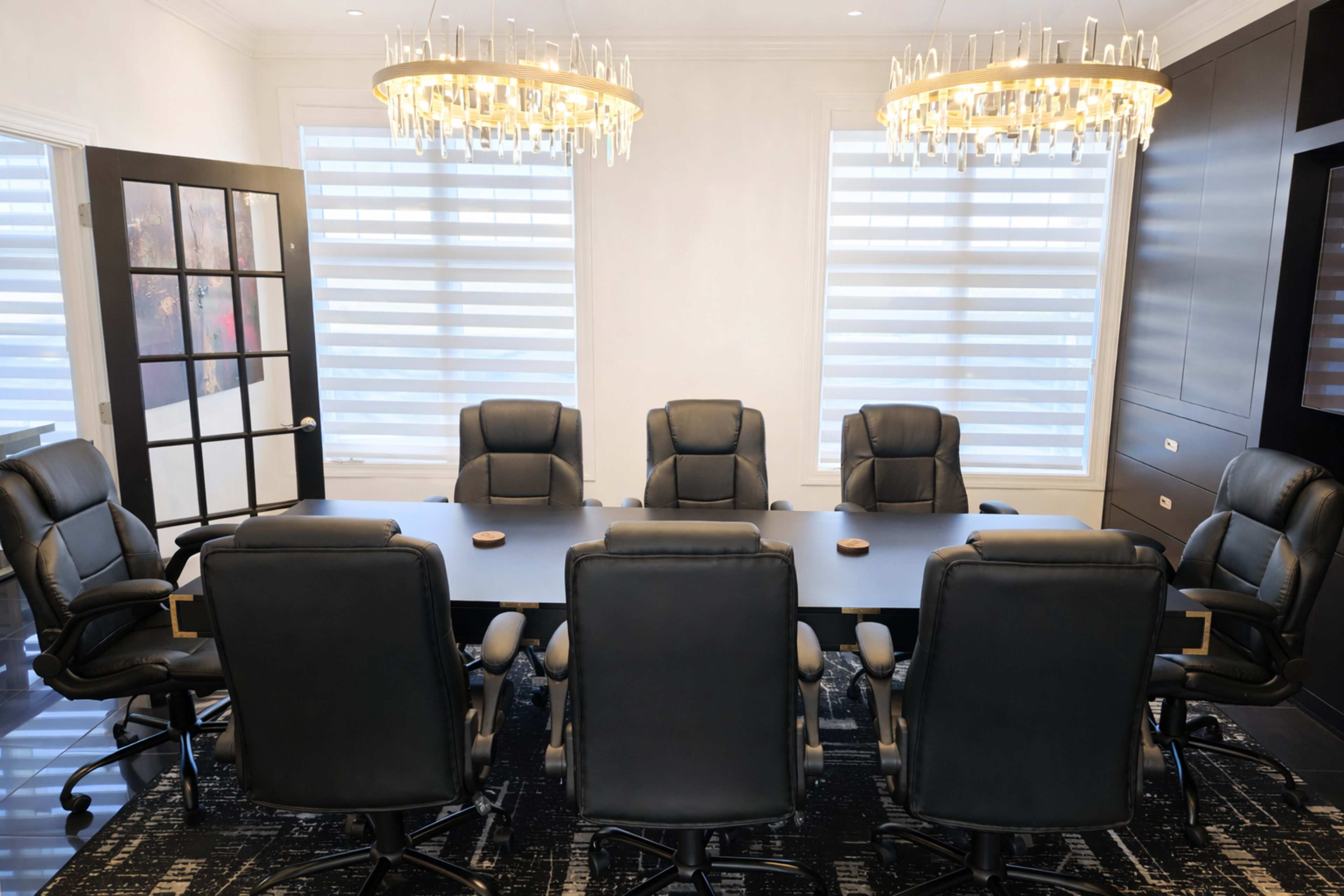 A conference room features a large black table surrounded by eight black swivel chairs, with two modern chandeliers overhead.