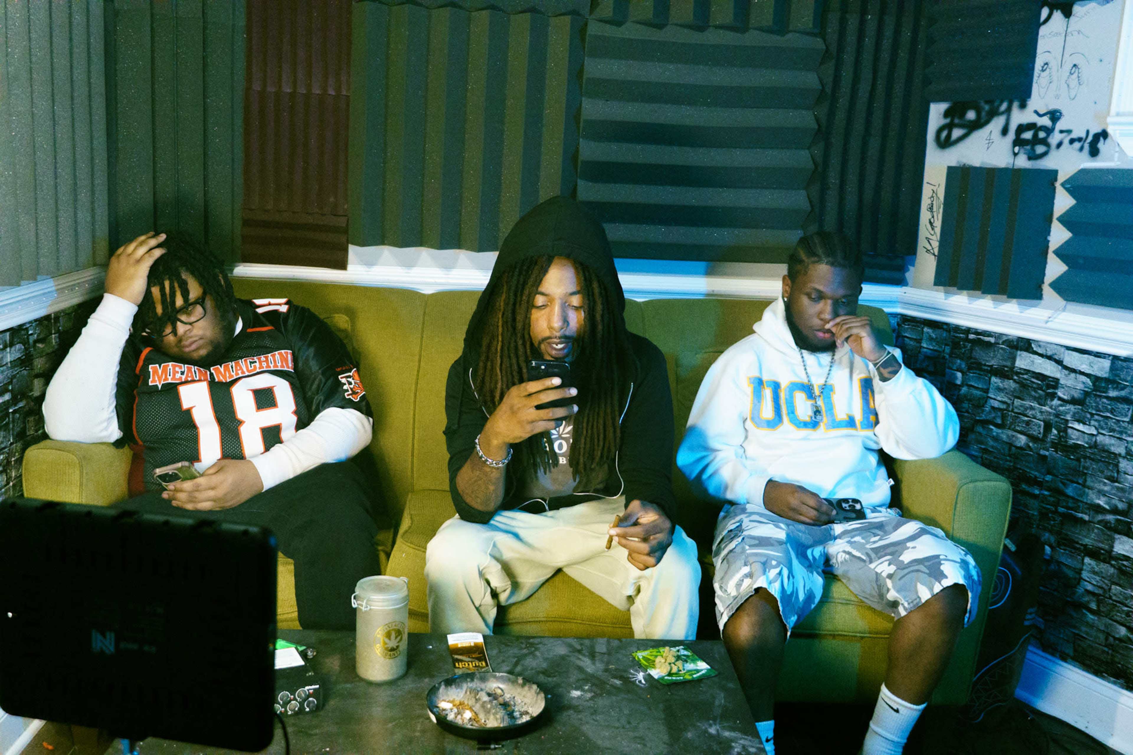 Three individuals sit on a green sofa in a room with soundproofing panels, each engaged with their phones, while various items are scattered on a low table in front of them.