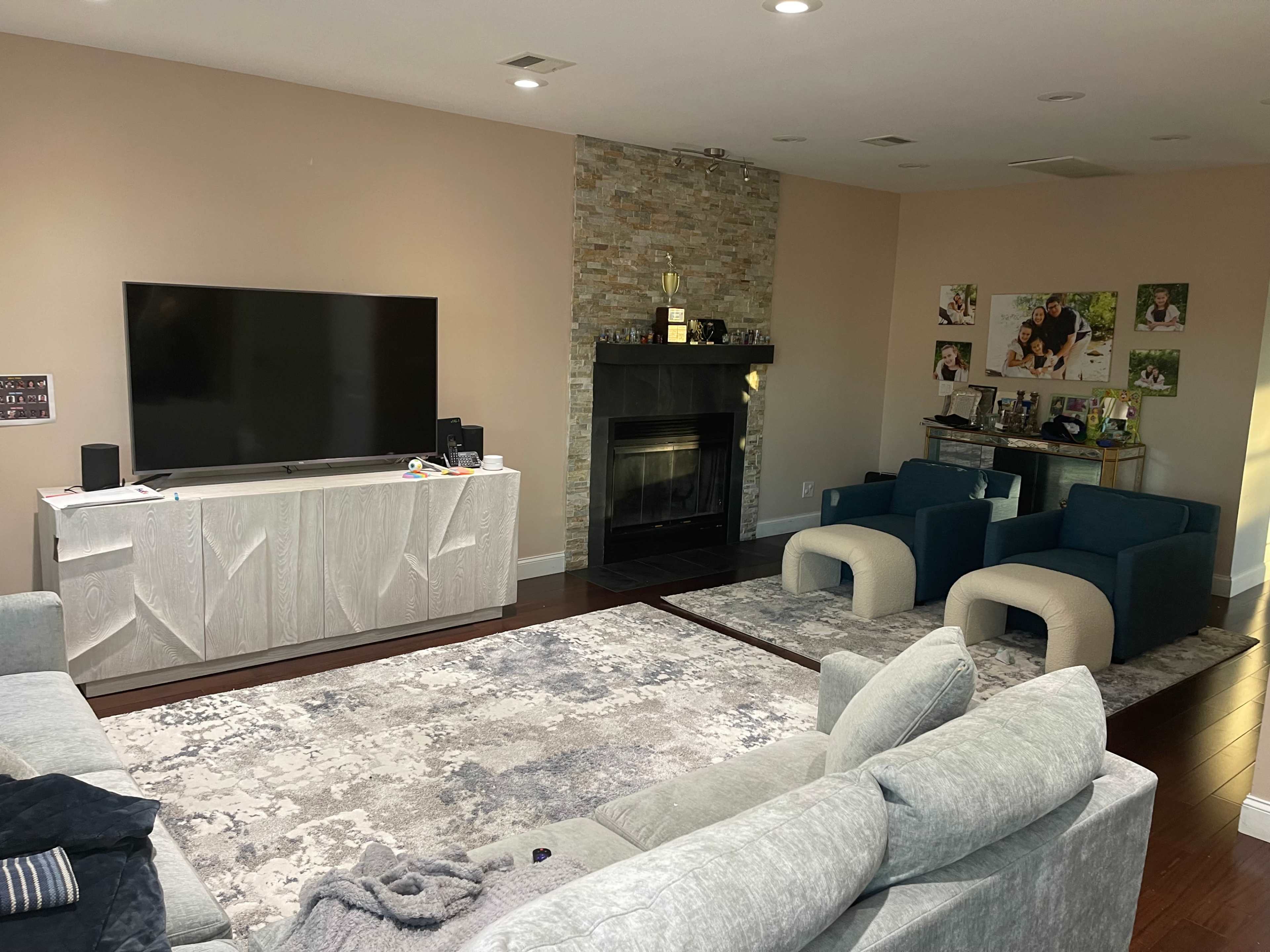 A modern living room features a large TV mounted on the wall, a stone fireplace, and two blue armchairs facing a light-colored sofa on a textured area rug.