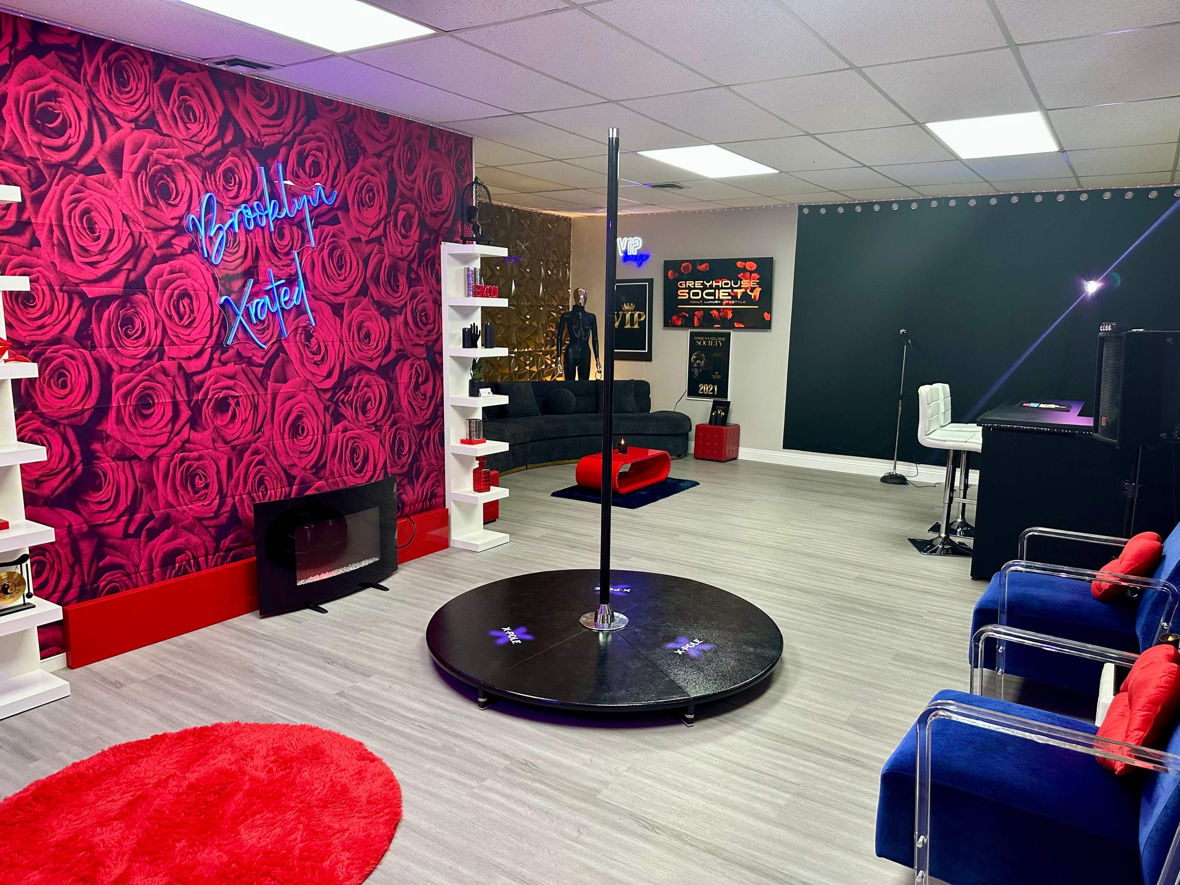 The image shows a colorful interior space featuring a black dance pole, a plush black sofa, a neon sign, and various decor elements against rose-patterned wallpaper.