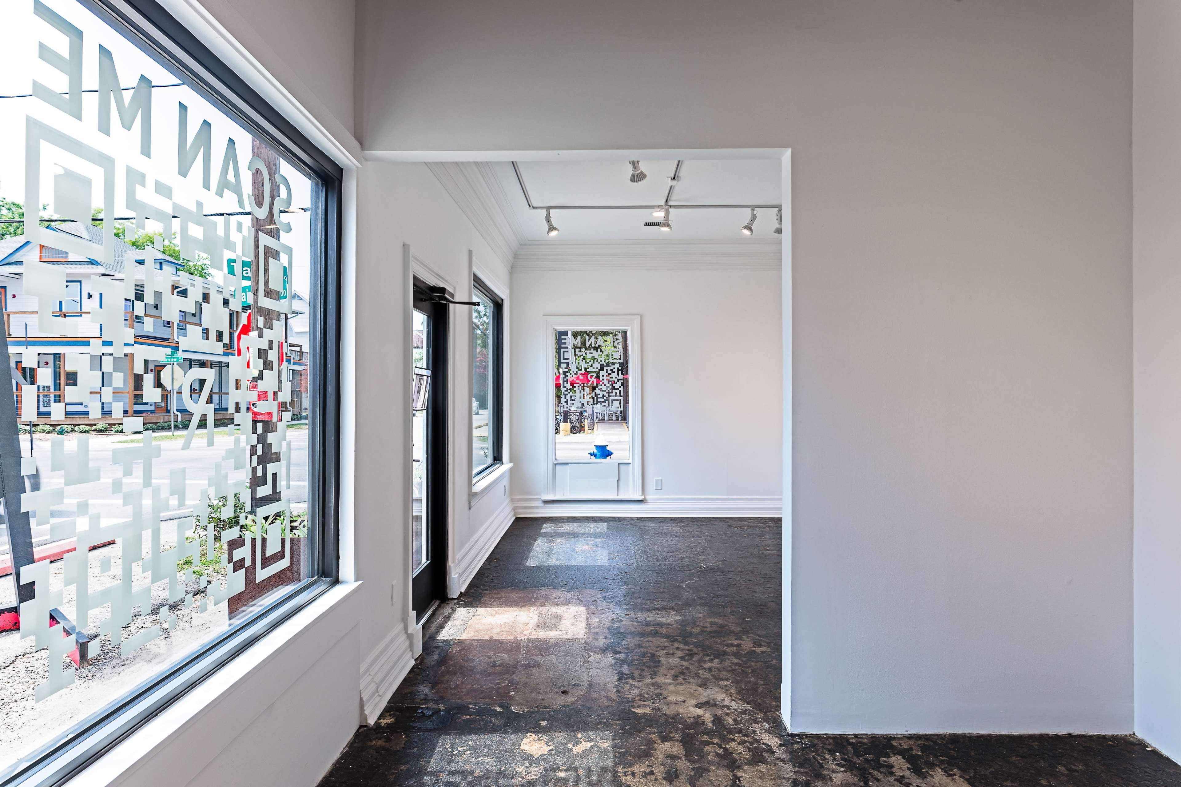 A spacious, well-lit art gallery with white walls and large windows facing an outdoor area.