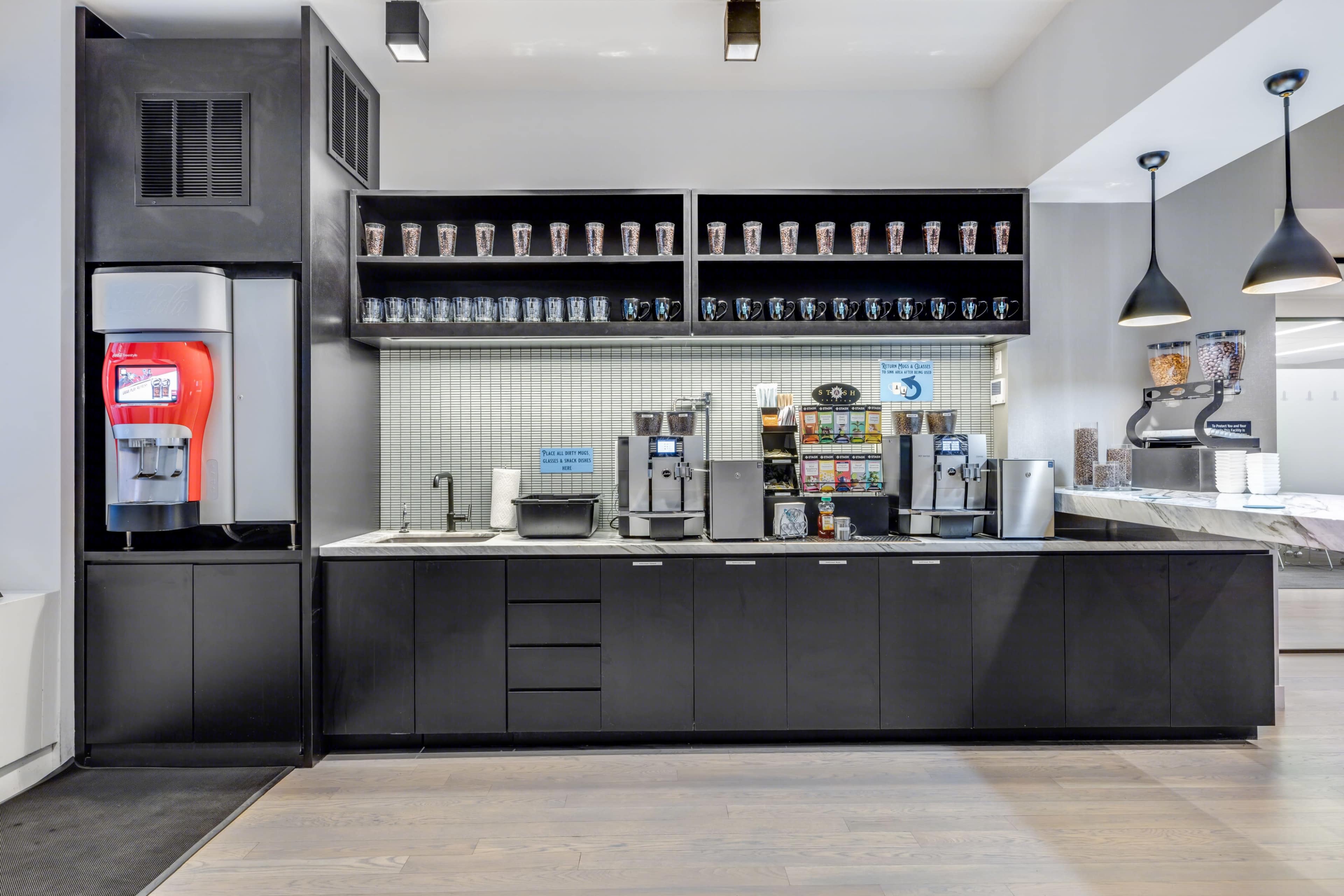 A modern beverage station with a drink dispenser, coffee machines, and shelves holding various cups in a sleek, black cabinetry.