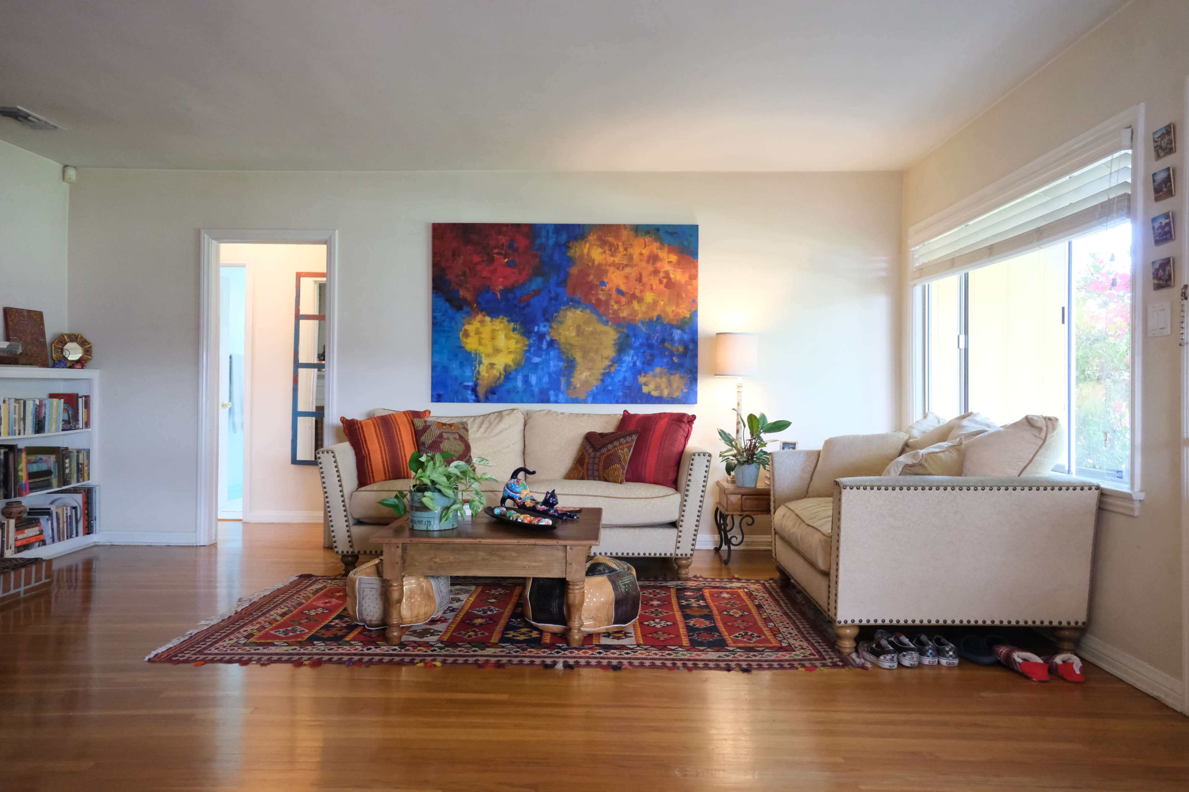 A living room features a beige sofa and armchair, a wooden coffee table, a large colorful world map painting on the wall, and a patterned rug.
