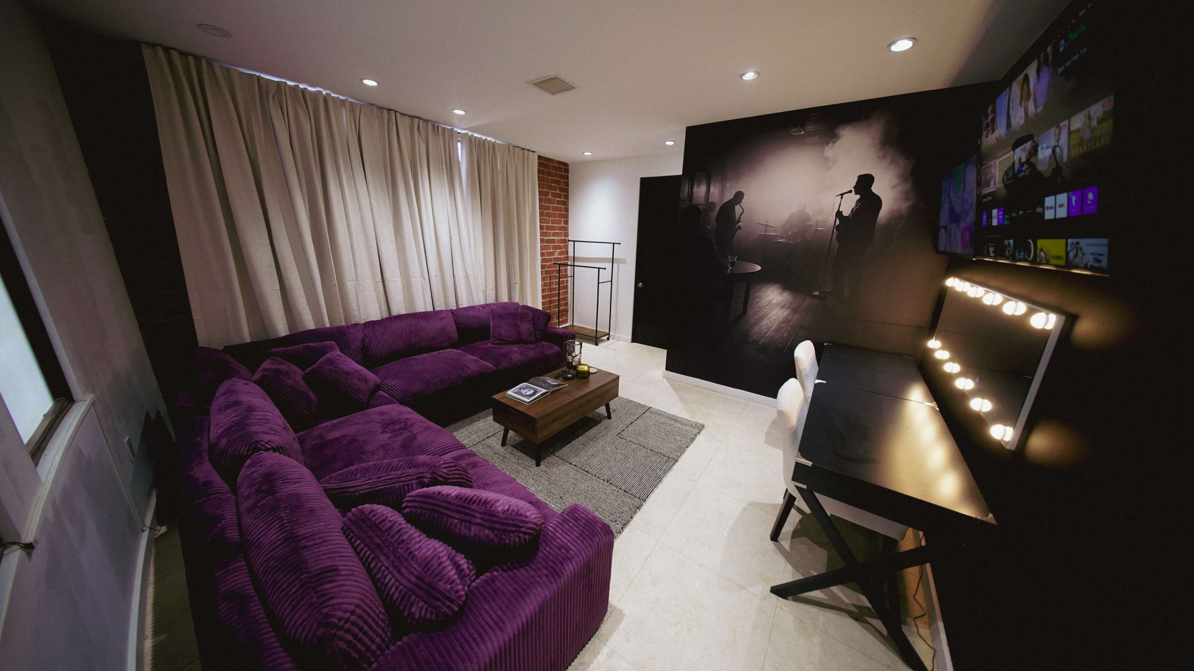The image shows a modern living room featuring a purple sectional sofa, a coffee table, and a desk with lighting, complemented by a wall with a black and white mural and curtains.