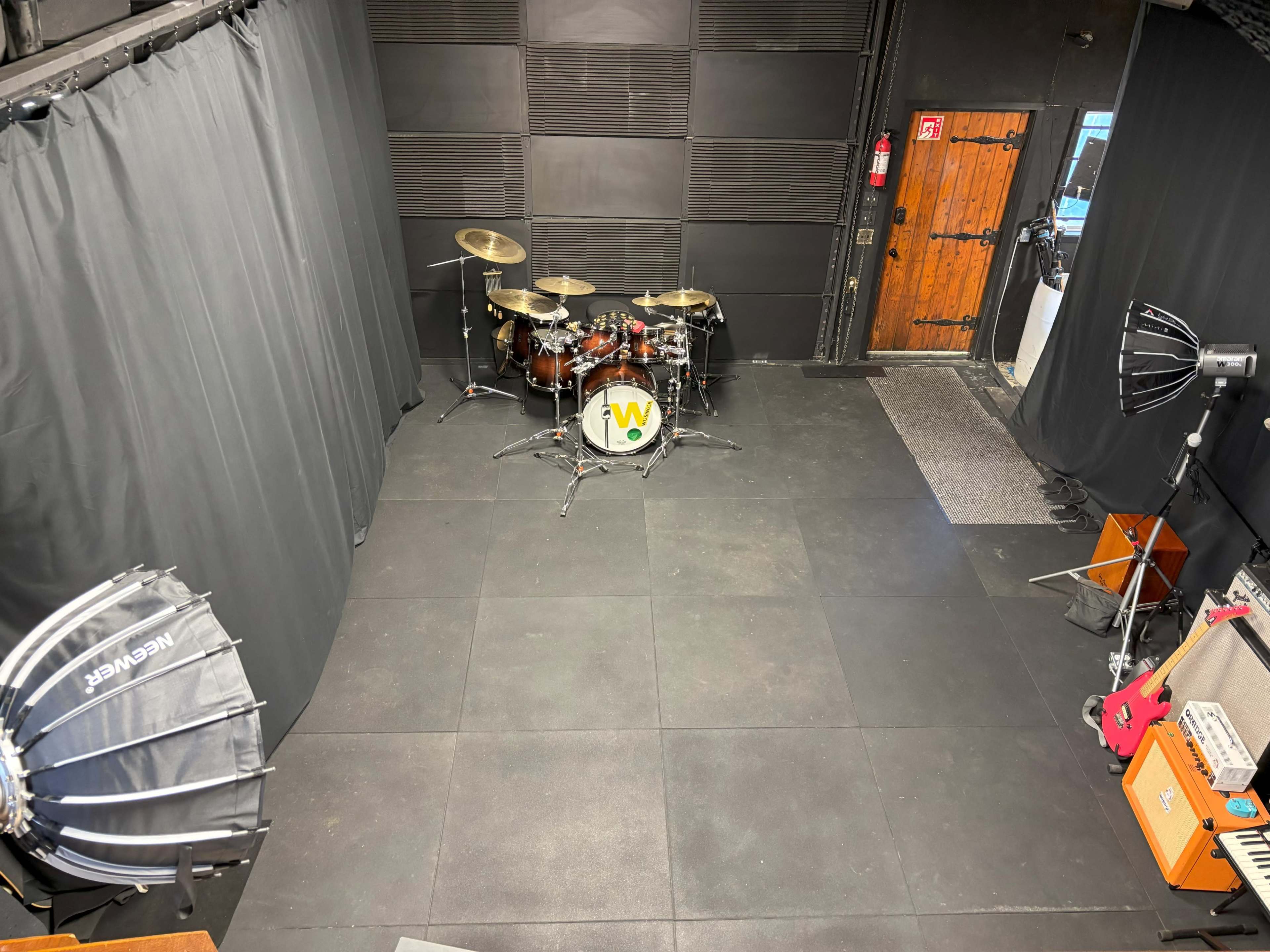 The image shows a music rehearsal space with a drum set in the center, surrounded by black curtains and various lighting equipment positioned around the room.