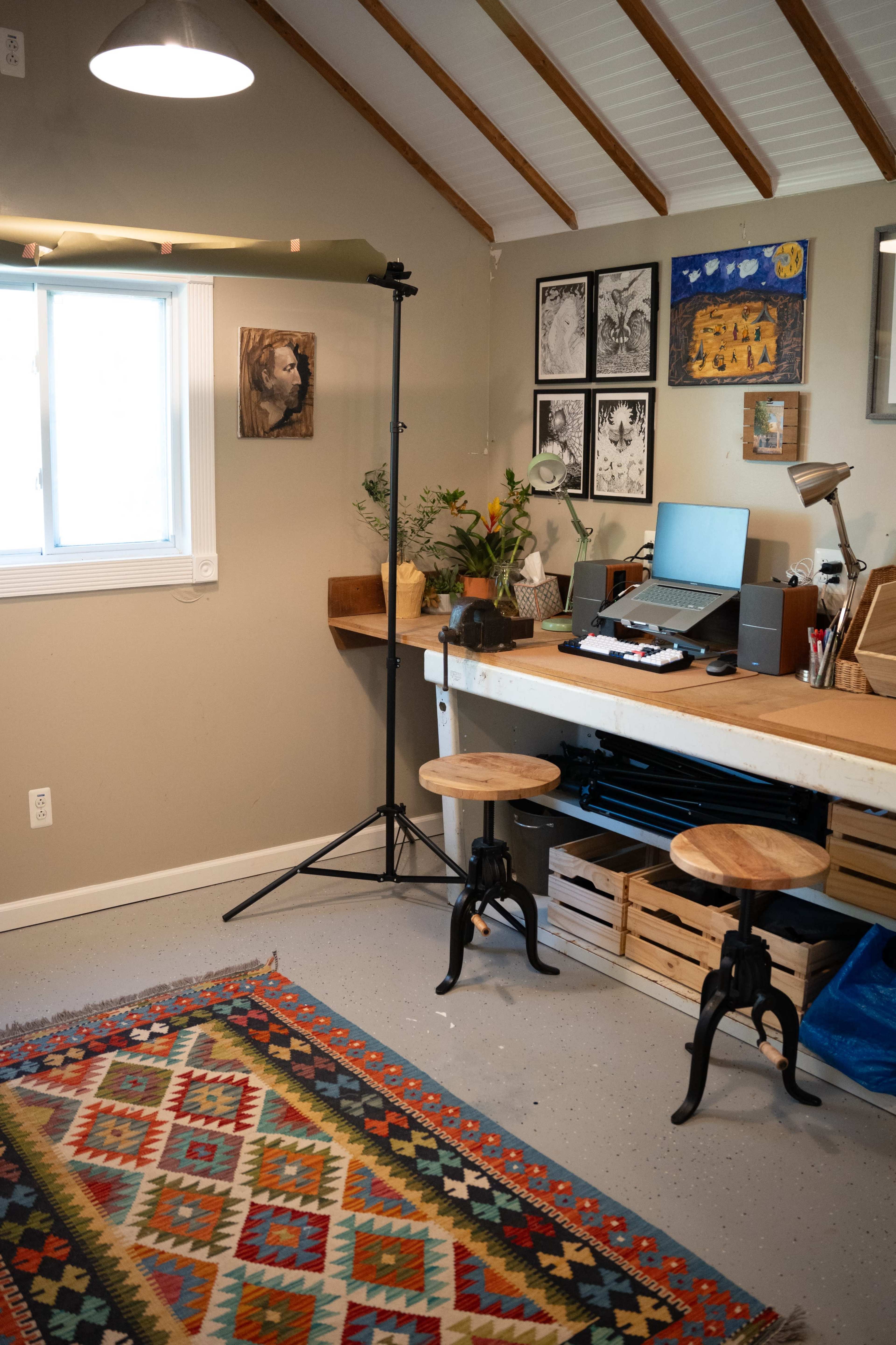 The image shows an artist's workspace with a large wooden table, two stools, a computer, and a light stand, surrounded by framed artwork on the walls.