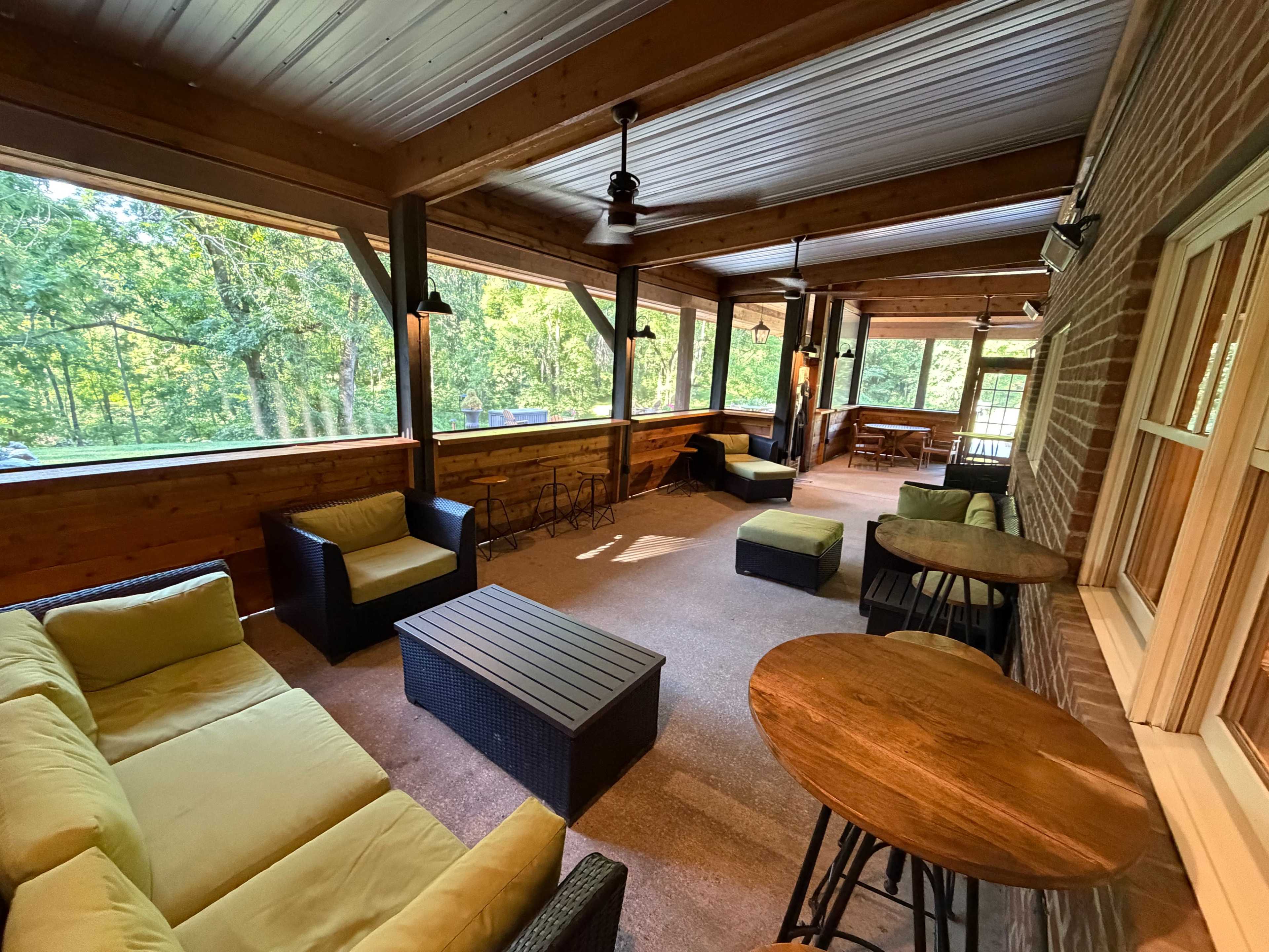 The image shows a screened-in porch with several seating areas, including a sectional sofa and round tables, overlooking a green wooded area.
