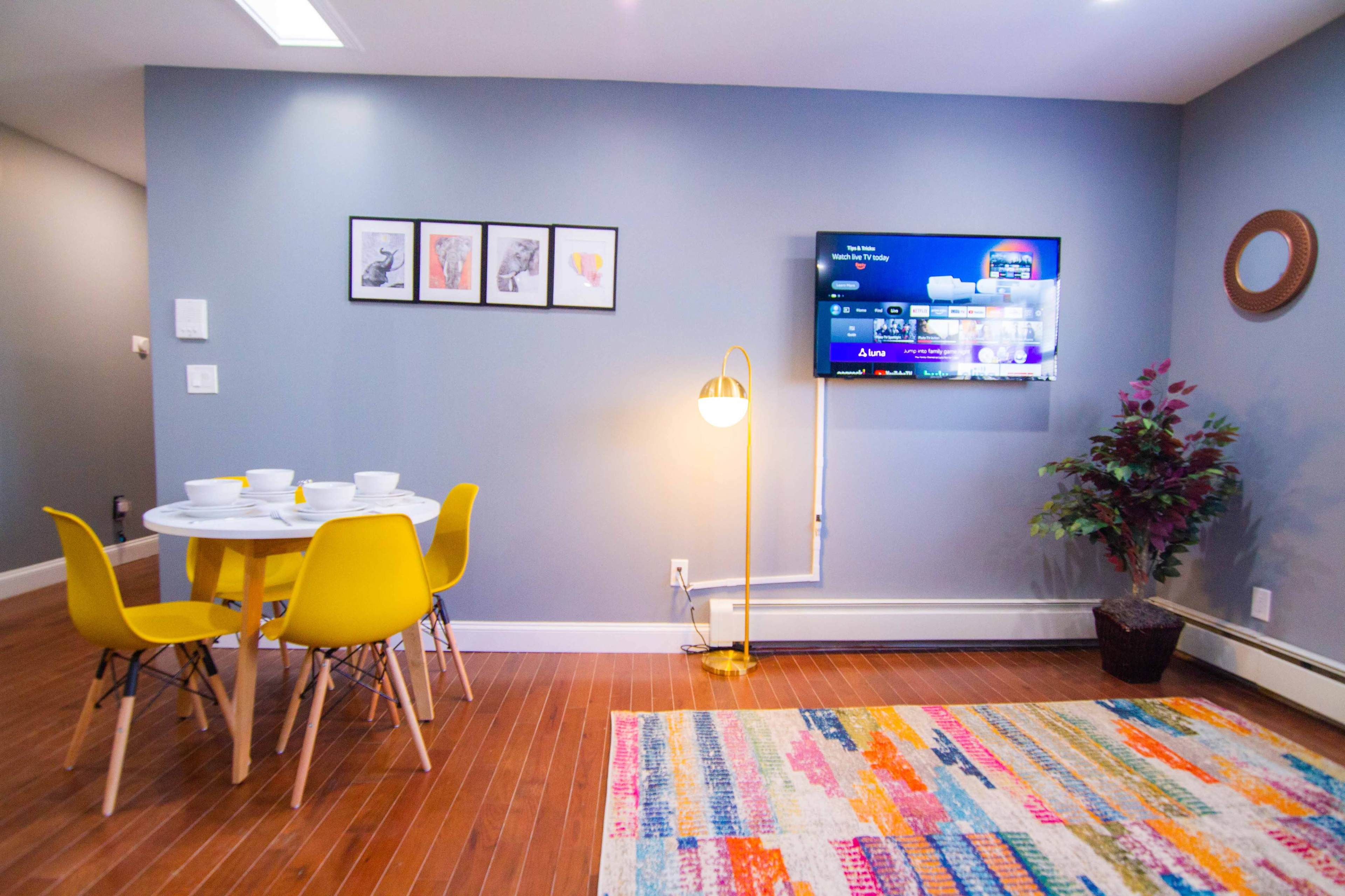 A modern living space features a round table with yellow chairs, a wall-mounted TV, framed artwork, and a decorative plant beside a colorful area rug.