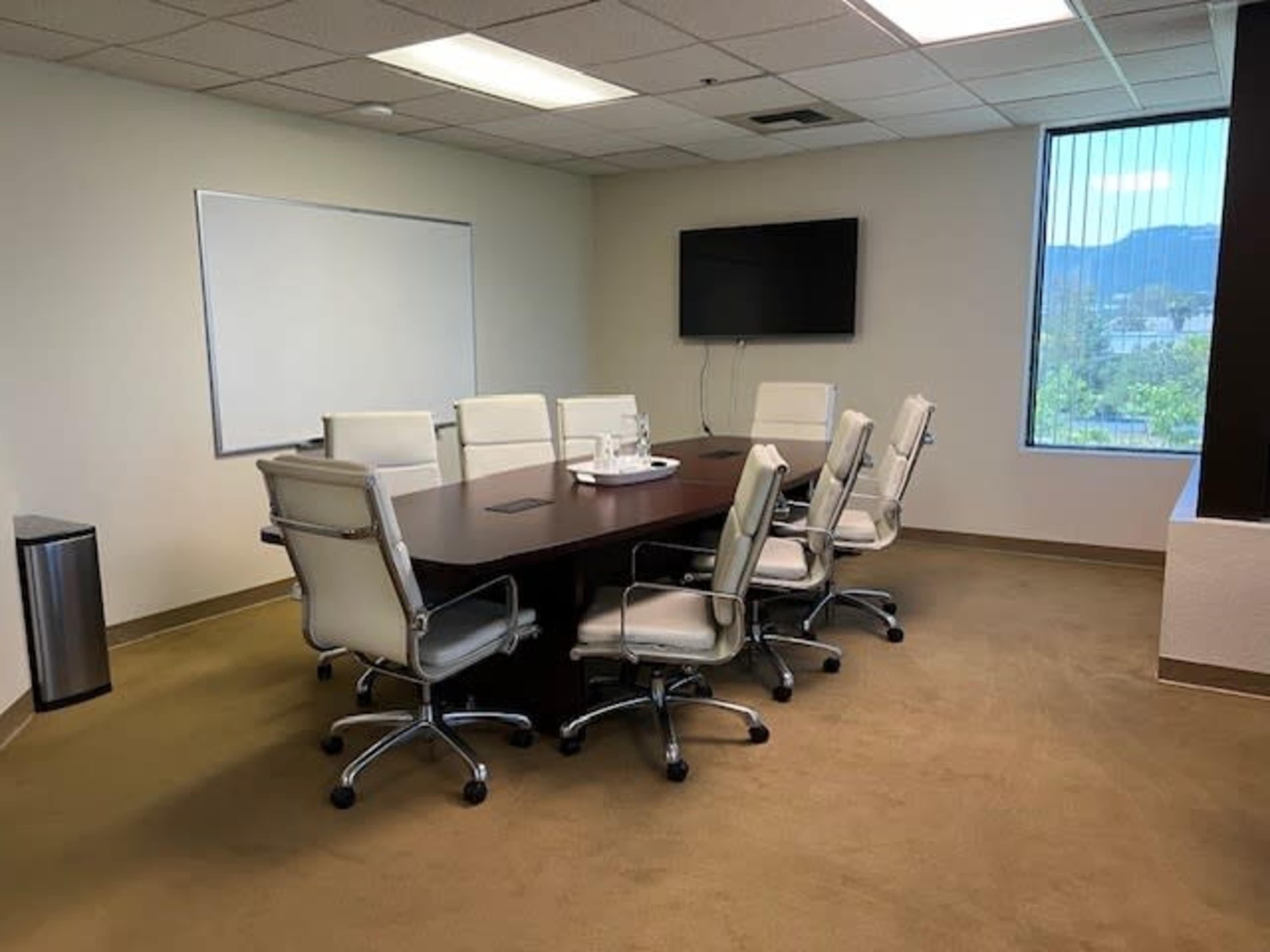 A conference room features a large wooden table surrounded by eight white leather chairs, with a wall-mounted television and a whiteboard visible in the background.