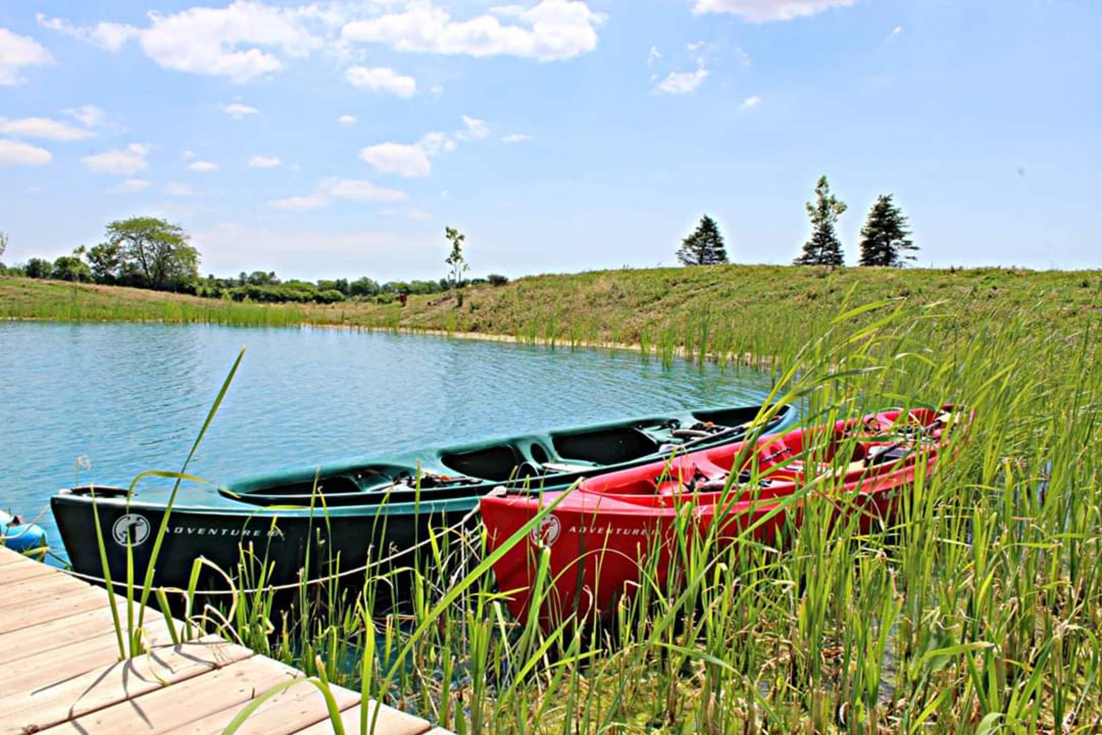 Two kayaks, one green and one red, are tied to a wooden dock beside a calm, blue lake surrounded by tall grass and trees.