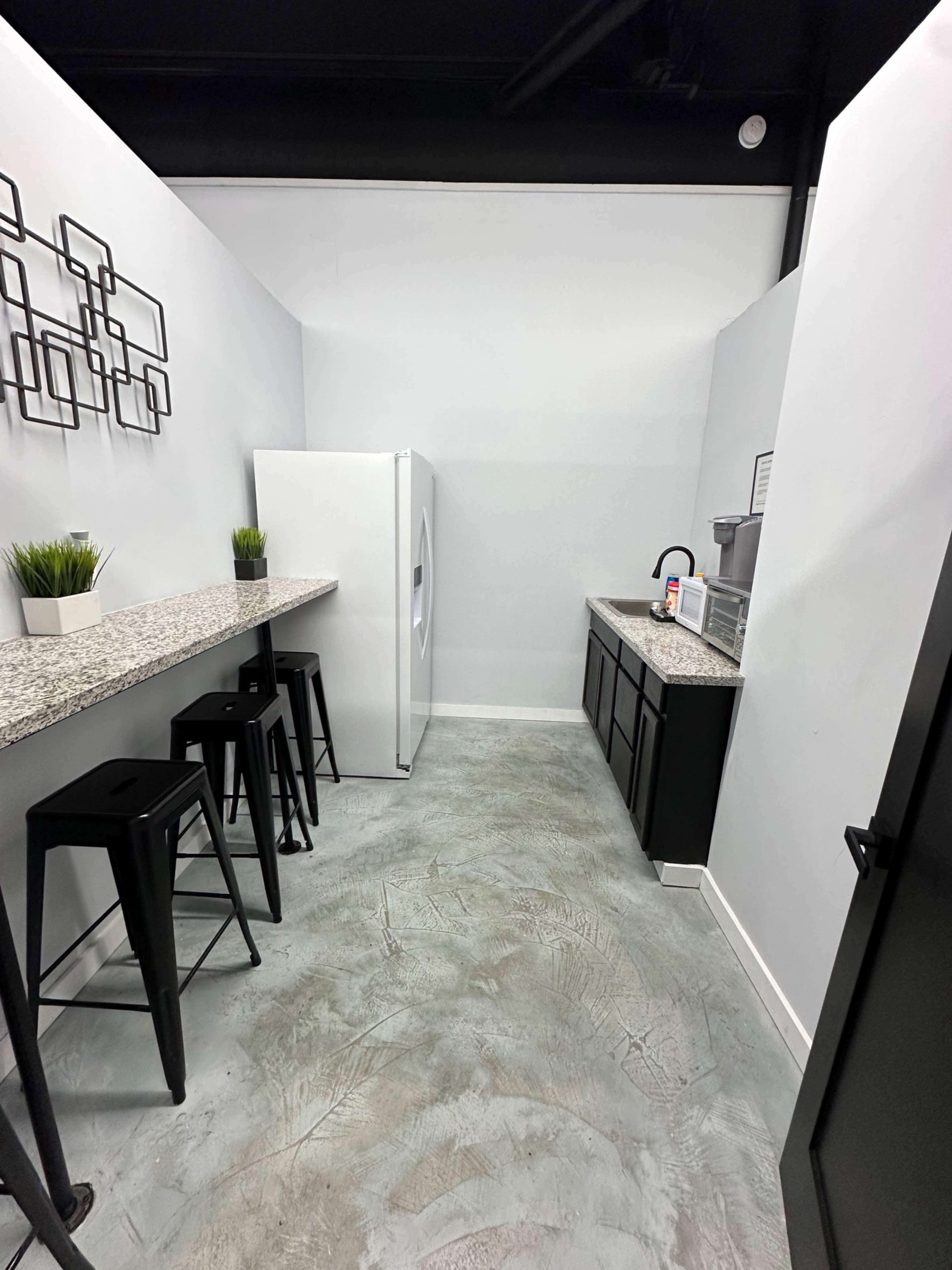 The image shows a minimalist kitchen area featuring a white refrigerator, dark cabinets, a countertop with stools, and a simple wall decor.