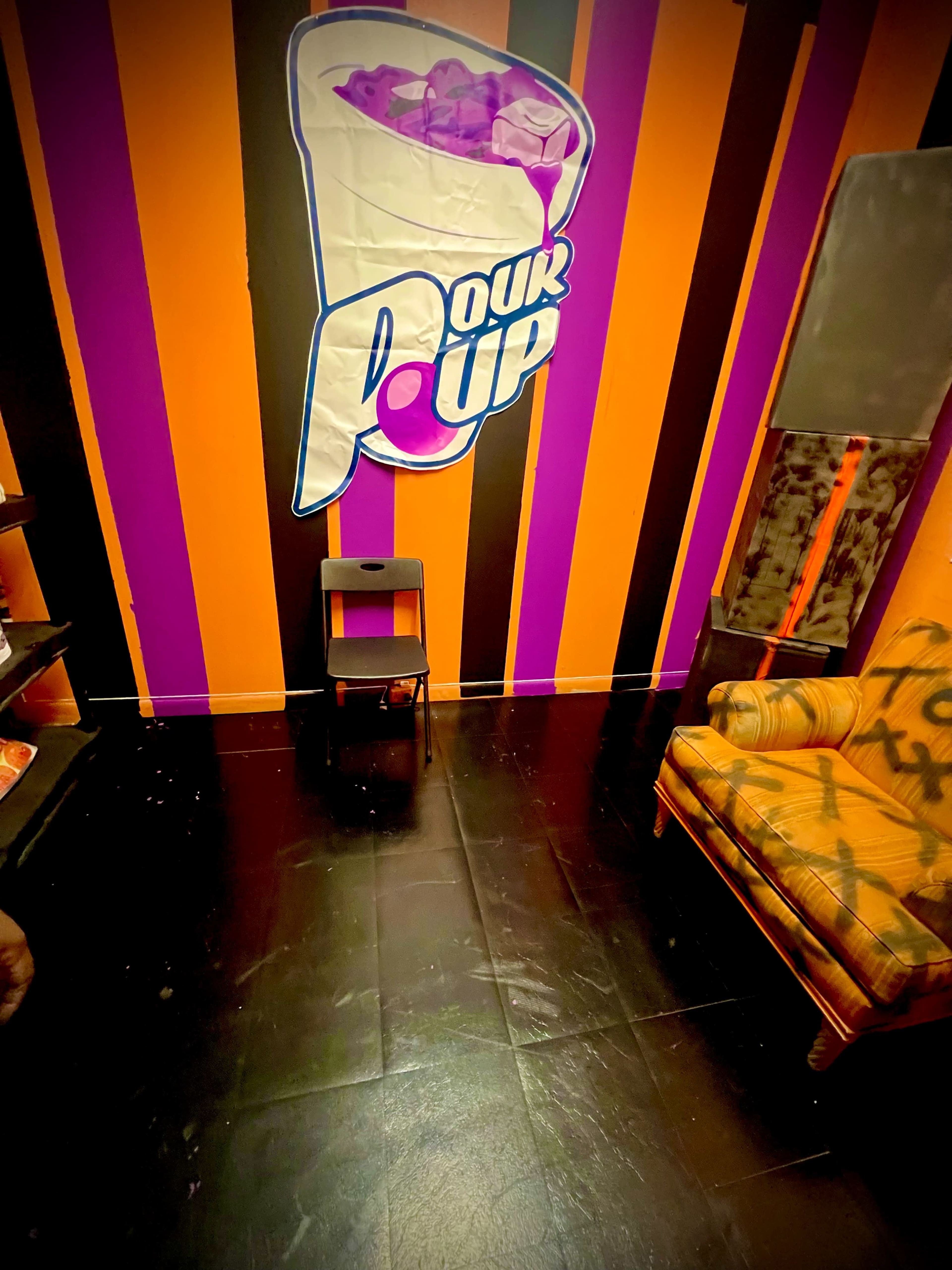 The image shows a room with orange and purple striped walls, a graphic of a drink on one wall, a patterned couch, and a small black stool.